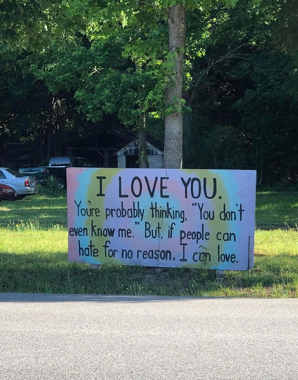 A sign in a park with pink, yellow, and blue paint with text over it. The text reads: "I LOVE YOU. You're probably thinking, 'You don't even know me.' But, if people can hate for no reason, I can love.