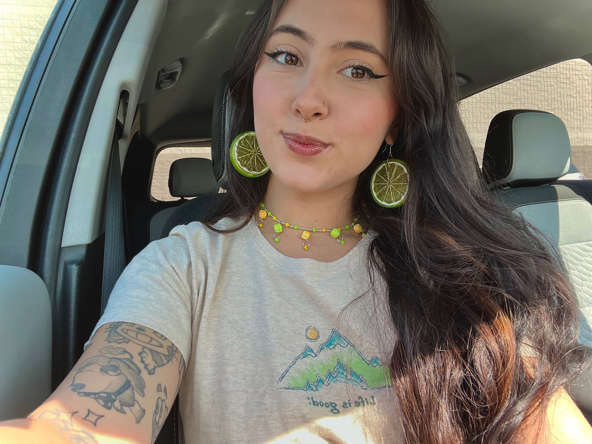 a picture of lorf, she is white and she has long dark hair and dark eyebrows. she has tattoos around her left upper arm. they are wearing green lime large dangly earrings and a yellow and green necklace they made. she is in a car.