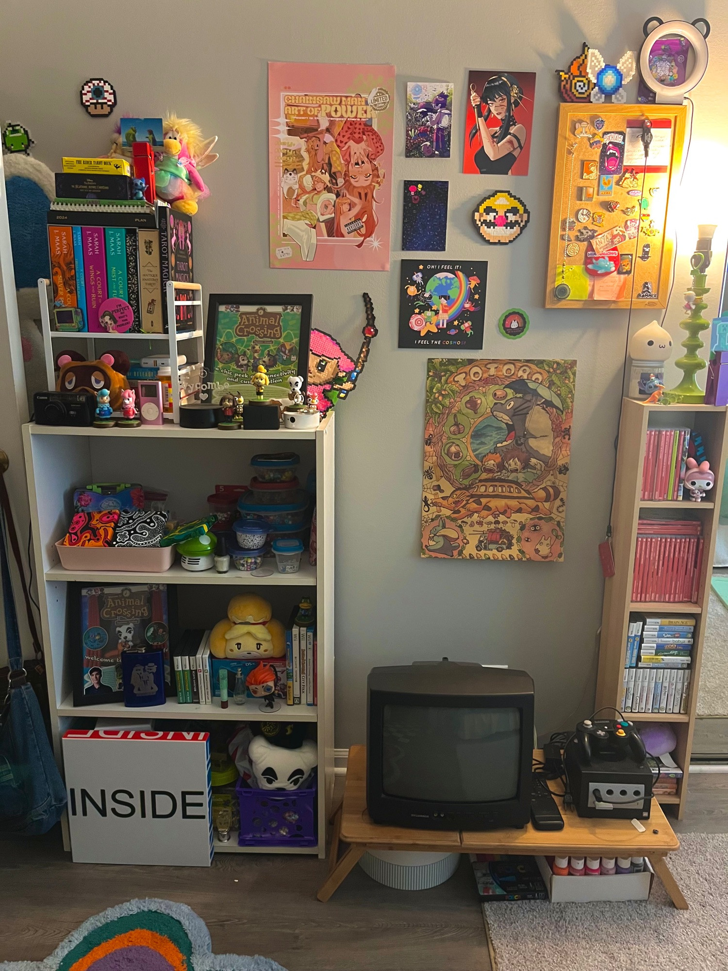 a picture of lorfs room. to the left there is a white shelf with various crafts and animal crossing merchandise scattered about. on the walls there are posters, some notably featuring totoro, yor forger, power from chainsaw man, and a katamari damacy art print. below the posters and to the right of the shelf, there is a small box tv and a gamecube. to the right is some of lorfs switch game collection, and some of their ds games. a top that collection is a lamp. there is a small flower rug on the floor visible at the bottom.