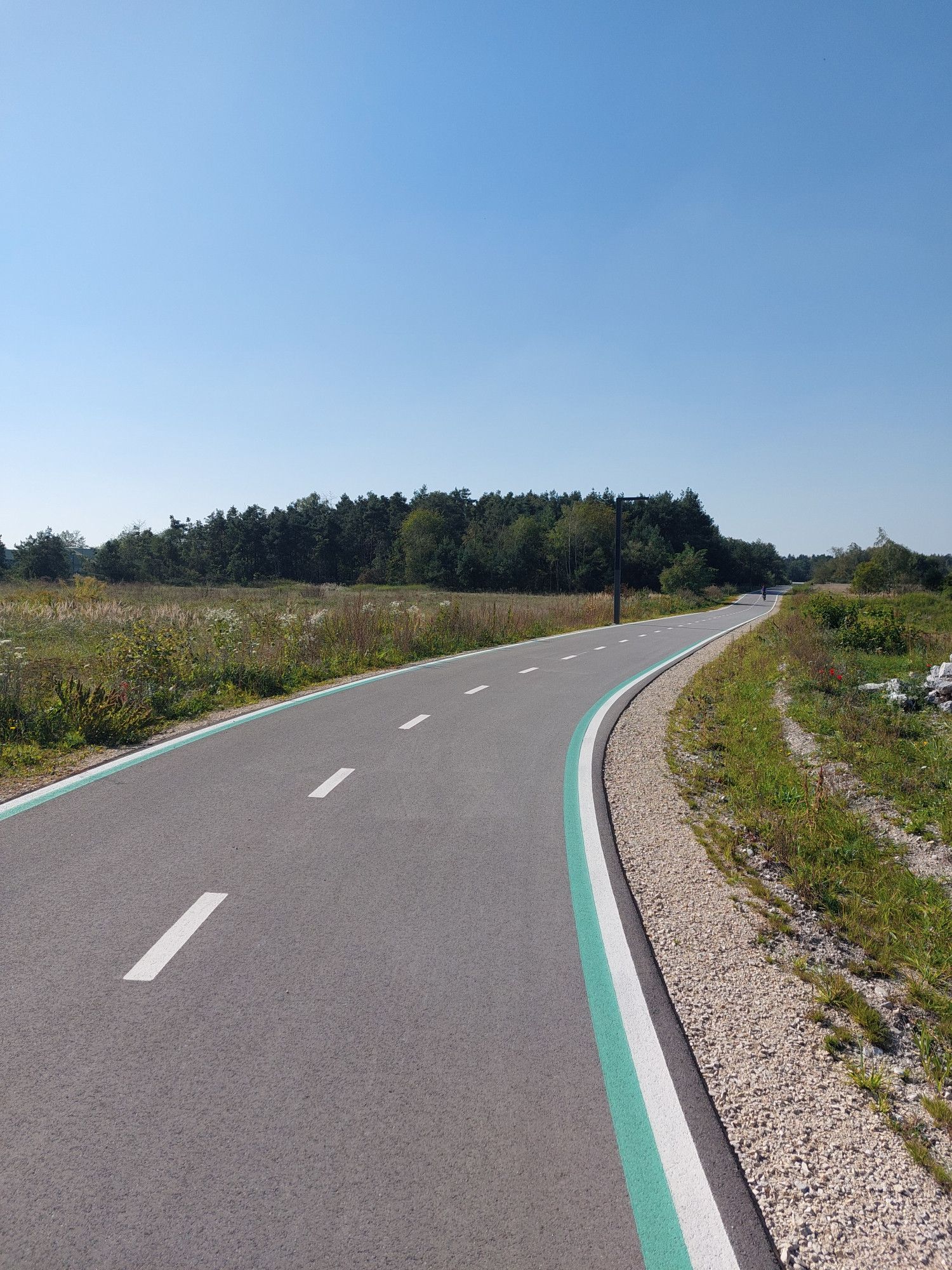 Wir sehen eine Straße, die uns in die lockende Ferne führt... aber dies ist doch keine Straße, die Seitenmarkierungen sind grün und weiß unter blauem Himmel. Rechts oben rast ein Radfahrer davon.