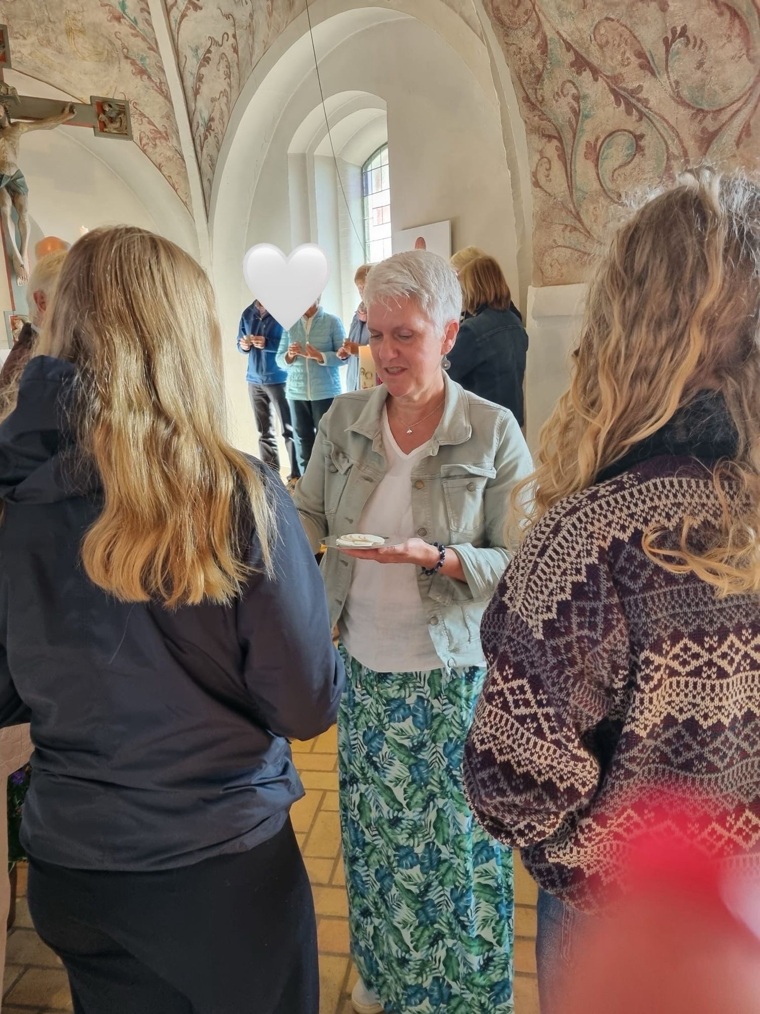 Frau mit hellen kurzen Haaren beim Austeilen des Brotes beim Abendmahl.  Sie steht schon zwei Personen mit langen blonden Haaren, die von hinten zu sehen sind im Altarraum einer Kirche.