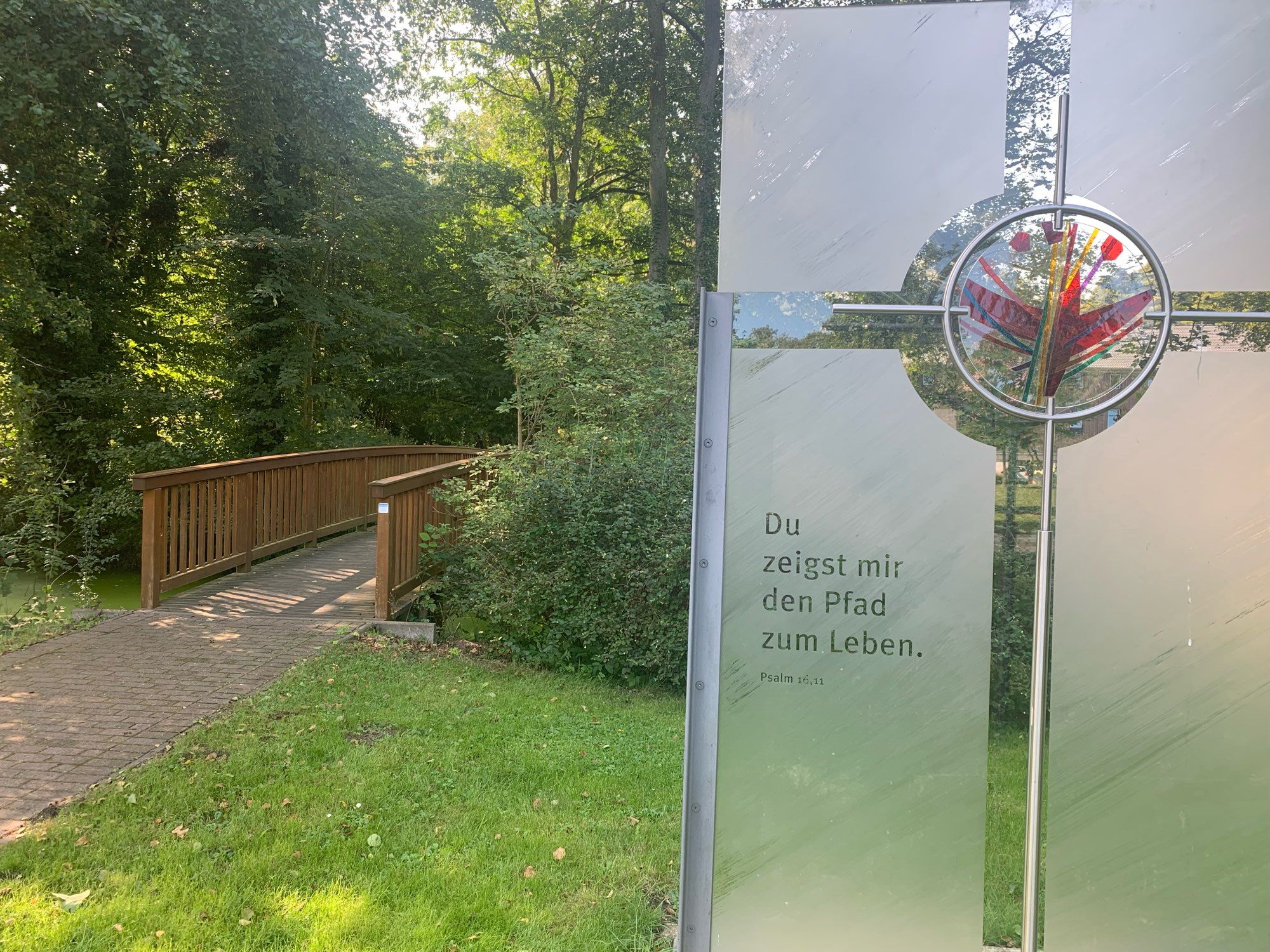 Du zeigst mir den Weg zum Leben. Psalm 16,11 auf Glas mit Kreuz. Links eine Holzbrücke.