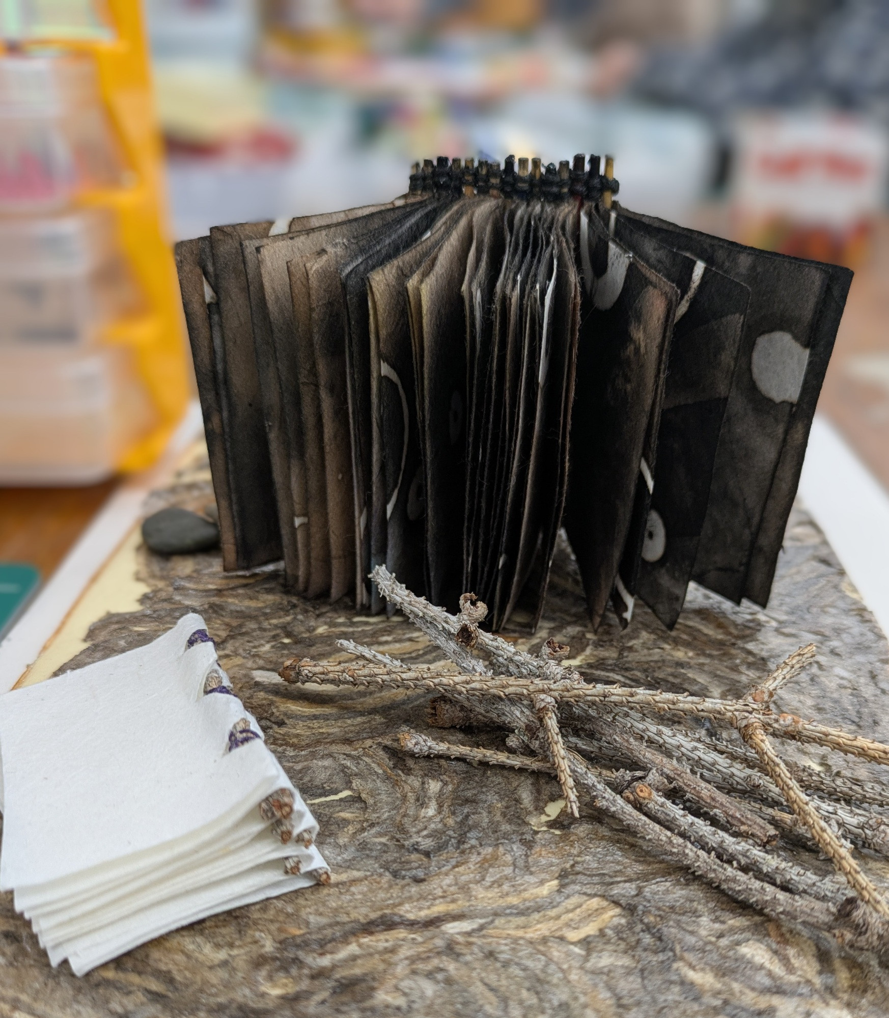 Two piano hinge books featuring sticks found on campus in the binding are shown next to a pile of pine twigs. The books are on top of a book cover made from hornets nest paper. 