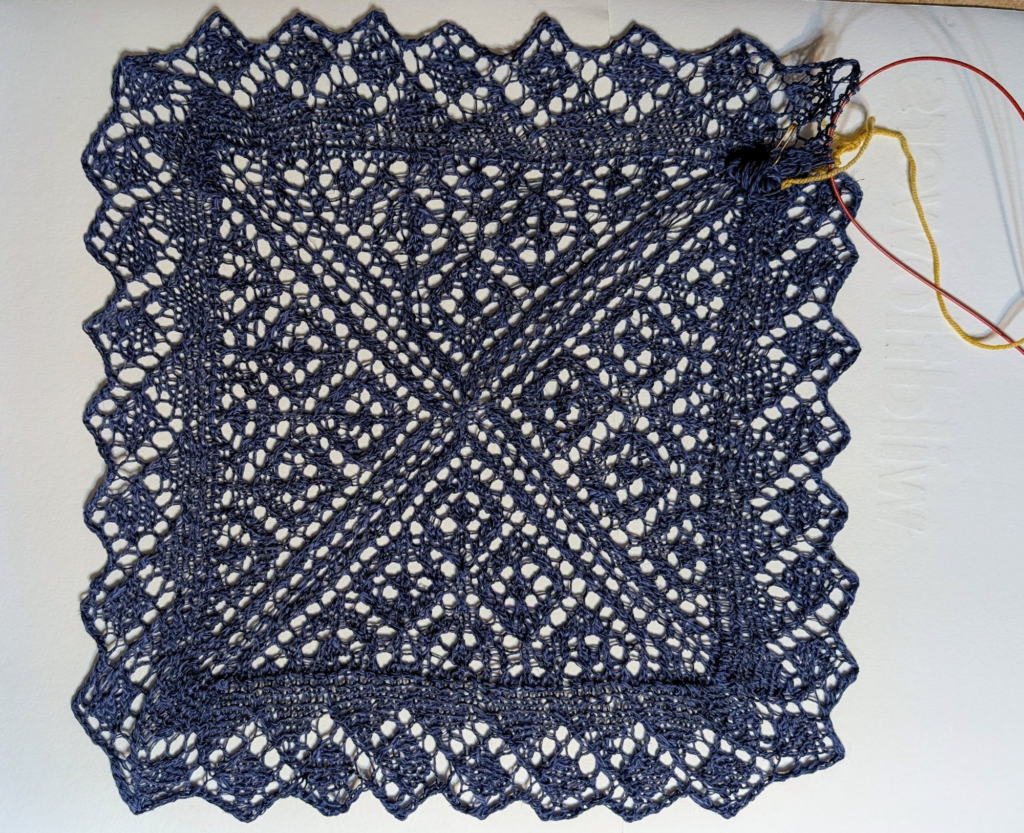 Dark blue knitted lace square. Diamond patterns form 4 triangles in the center and also appear in the lacey edging. 