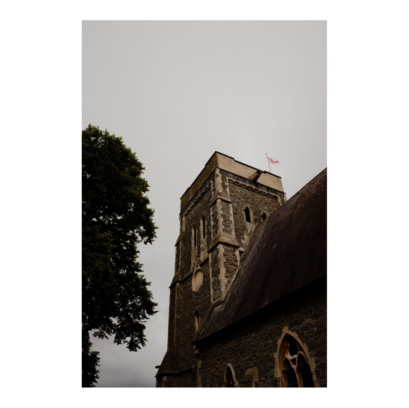 A square stone church tower against a gloomy sky. There is a flag of St. George flying from the tower.