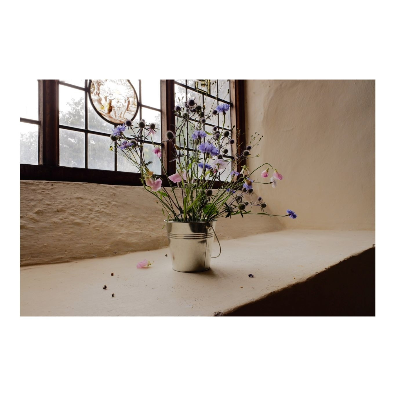 A vase of pink and purple flowers on the window ledge of a country church with stained glass behind.