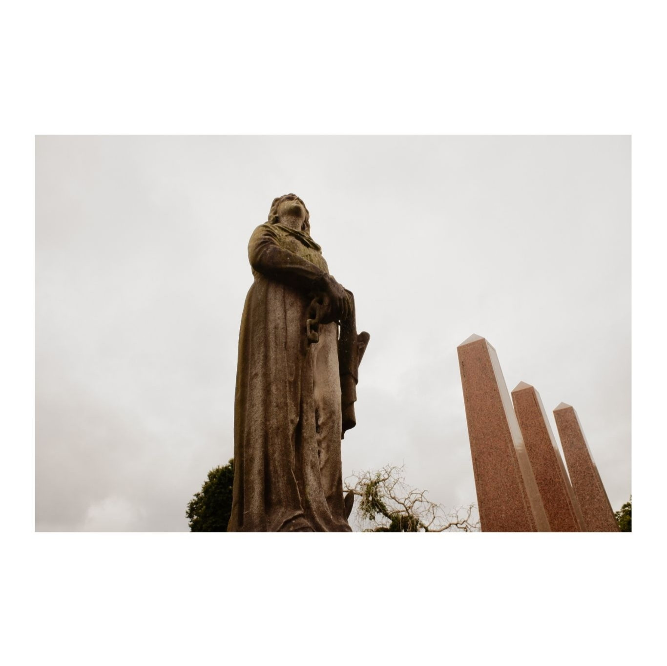 An imperious statue above a grave against a grey sky. Behind is a row of marble obelisks.