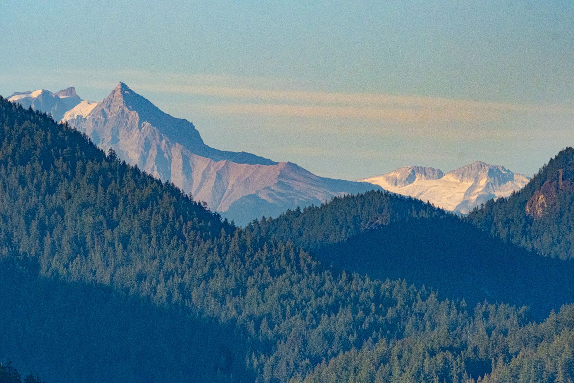 Smaller mountains in the foreground, densely forested. Behind them, rocky peaks with not-terribly-white snow. Above, a hazy blue sky. 