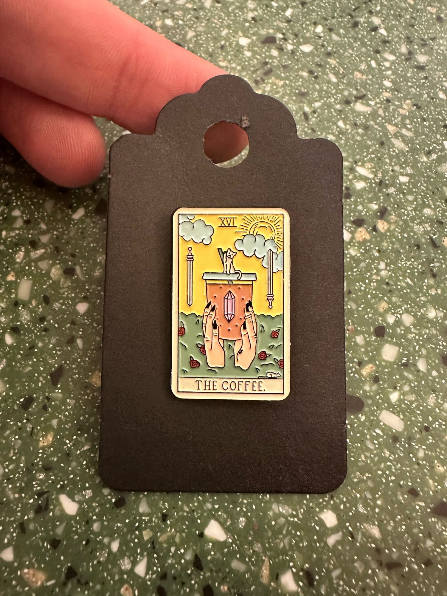 An enamel pin designed to look like a classically illustrated tarot card. It features two hands holding a to-go cup of coffee. The cup has a pinkish crystal on its side, and a cat rests on top next to the straw. The Roman numerals for 16 are on the top of the image, on the bottom of the image it reads “the coffee”.