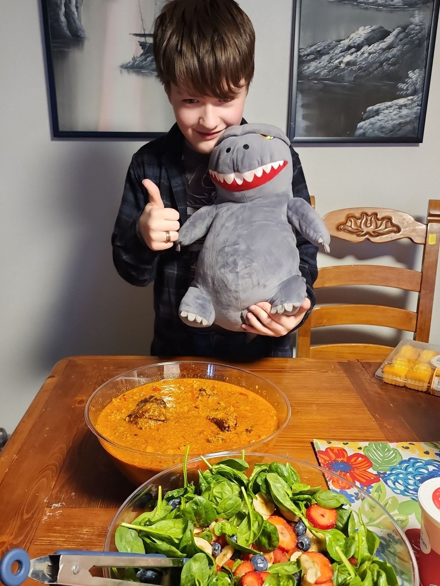 A person holding a **stuffed Godzilla toy** and giving a **thumbs up**, standing behind a table with a bowl of **salad** and chili dish. The background features black-and-white artworks depicting mountainous landscapes.