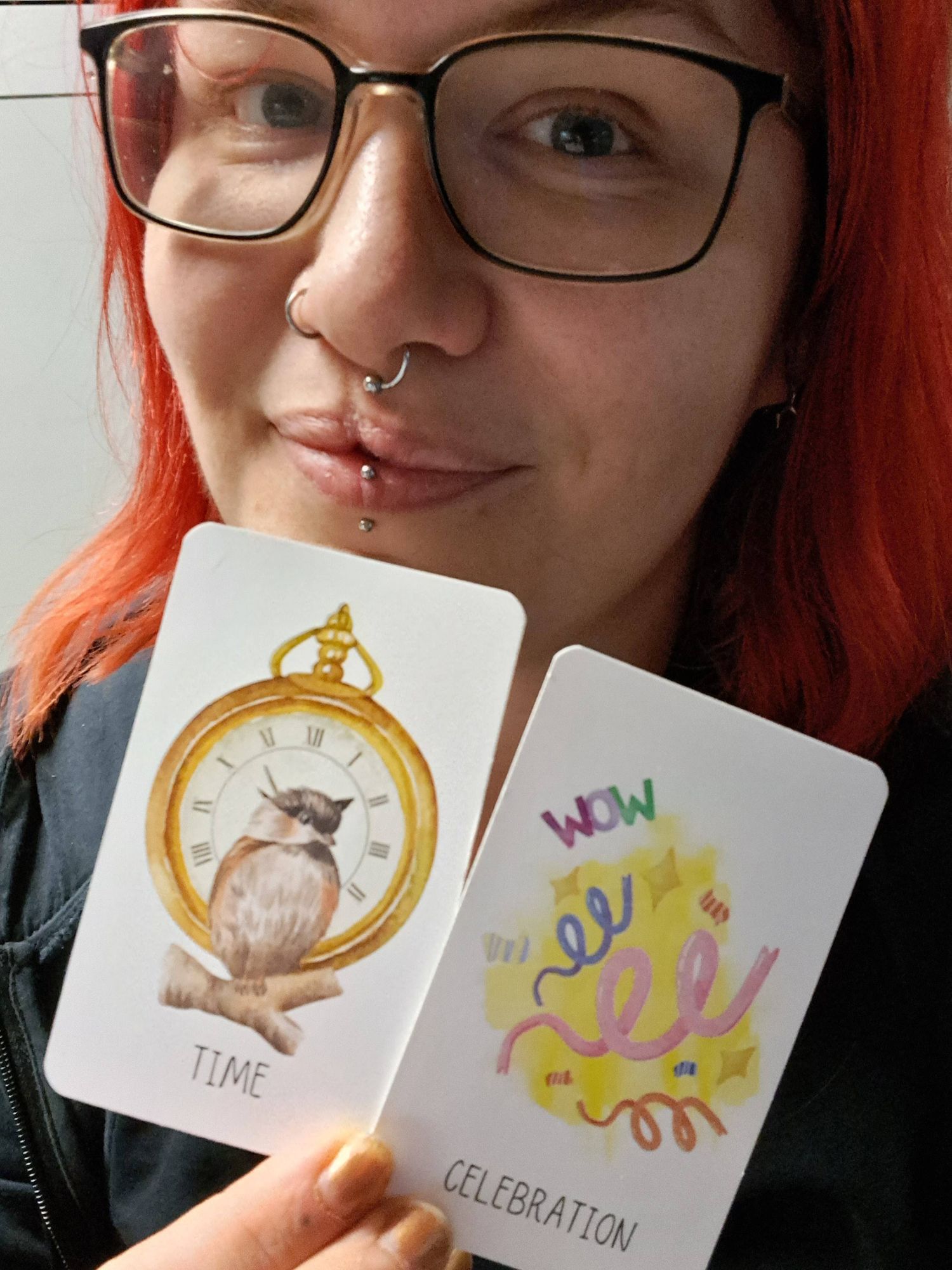 Pierced woman with red hair and big glasses is Holding two oracle cards which say time and Celebration. Time shows a clock and a sparrow and Celebration is showing confetti and the word WOW