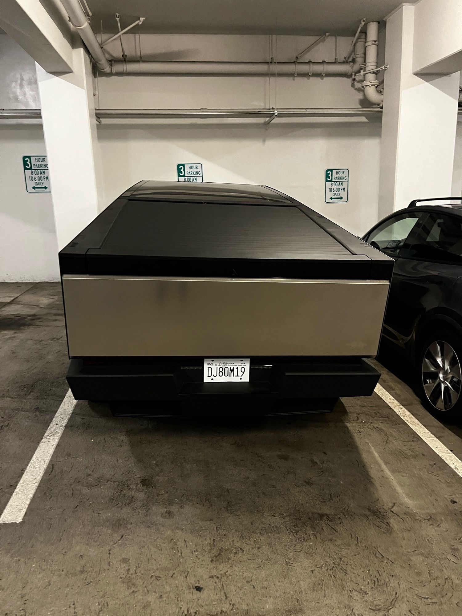 Cybertruck pictured from the rear, parked in a small parking spot in a public parking structure. Vehicle almost touches the lines on each side of the parking spot. The vehicle appears visually as a trapezoid over a rectangle and is, from this caption-writer’s perspective, exceptionally unappealing to look at.