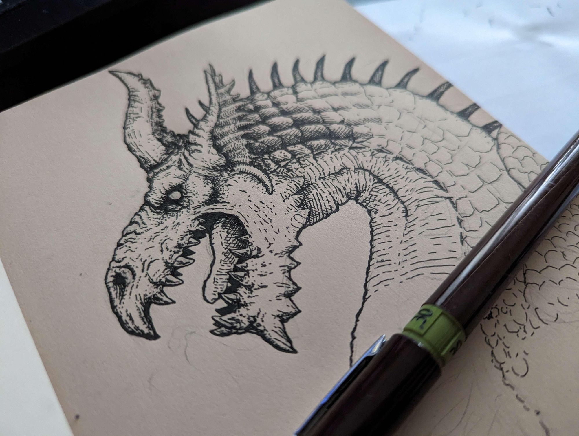 Picture of a sketchbook. You see a dragon portrait, made with ink, and an ink pen. The dragon is a character from the video game Dragon's Dogma: Grigori. 