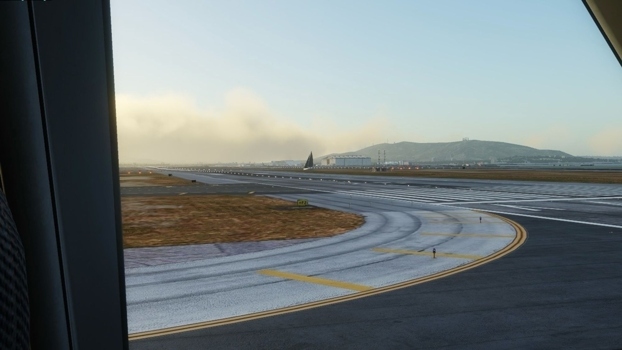 A XPlane 12 simulated view from an airplane cockpit window showing an airport runway with taxiways and lighting, set against a background of mountains and buildings. The sky is clear with some scattered clouds. There’s a sail boat just down the runway.