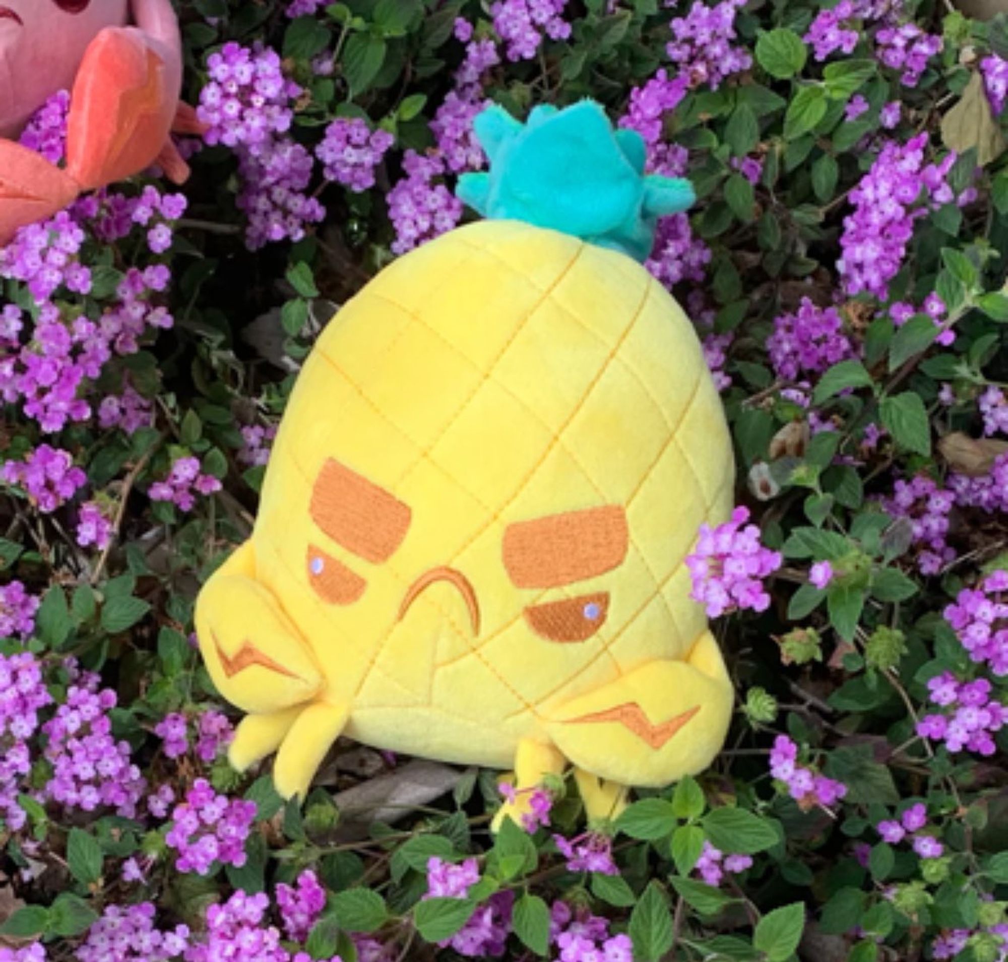 Photo of a yellow pineapple crab plush toy in a bush with purple flowers.