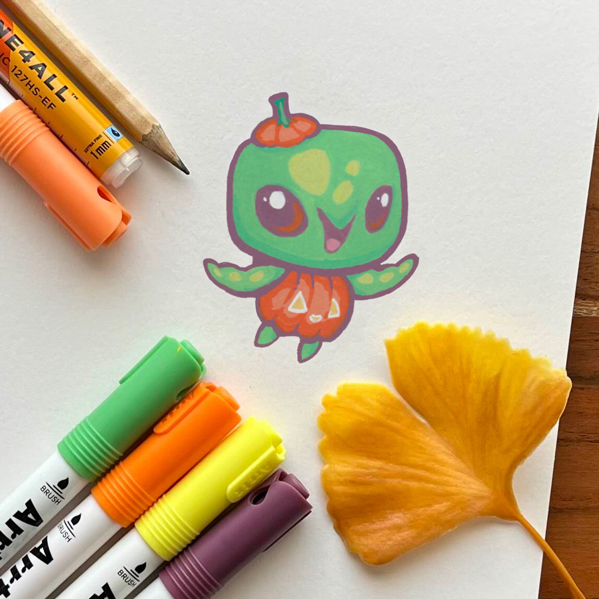 Photo of an illustration featuring a baby sea turtle character design. They are wearing a tiny jack o' lantern and the stem lid as a hat. The spot illustration is surrounded by markers and a yellow gingko leaf for seasonal flair.