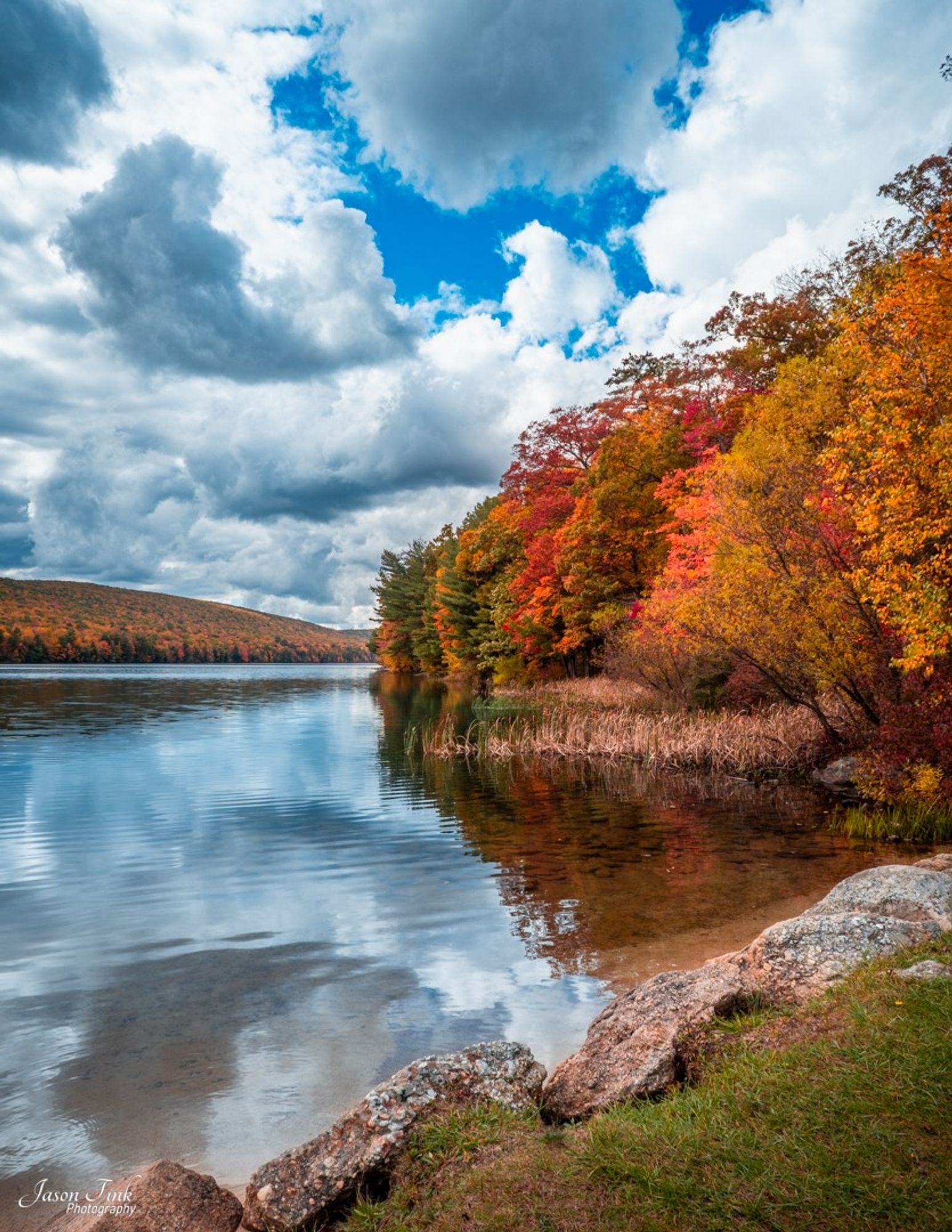 Vibrant autumn leaves reflecting in the tranquil waters of Mauch Chunk lake create a serene landscape. Fluffy white clouds dot the sky, adding to the picturesque Pocono Mountain scene set against a backdrop of a distant forest-covered hill.