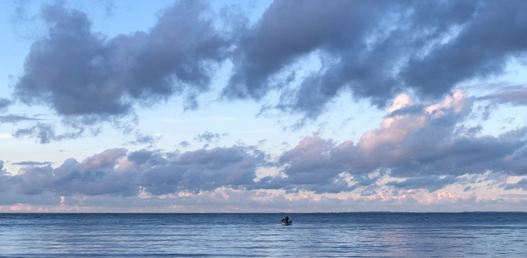 Approaching dusk. Pale blue sky with clouds filling most of the picture above a strip of calm sea with a fisherman in a small motor launch near the horizon.