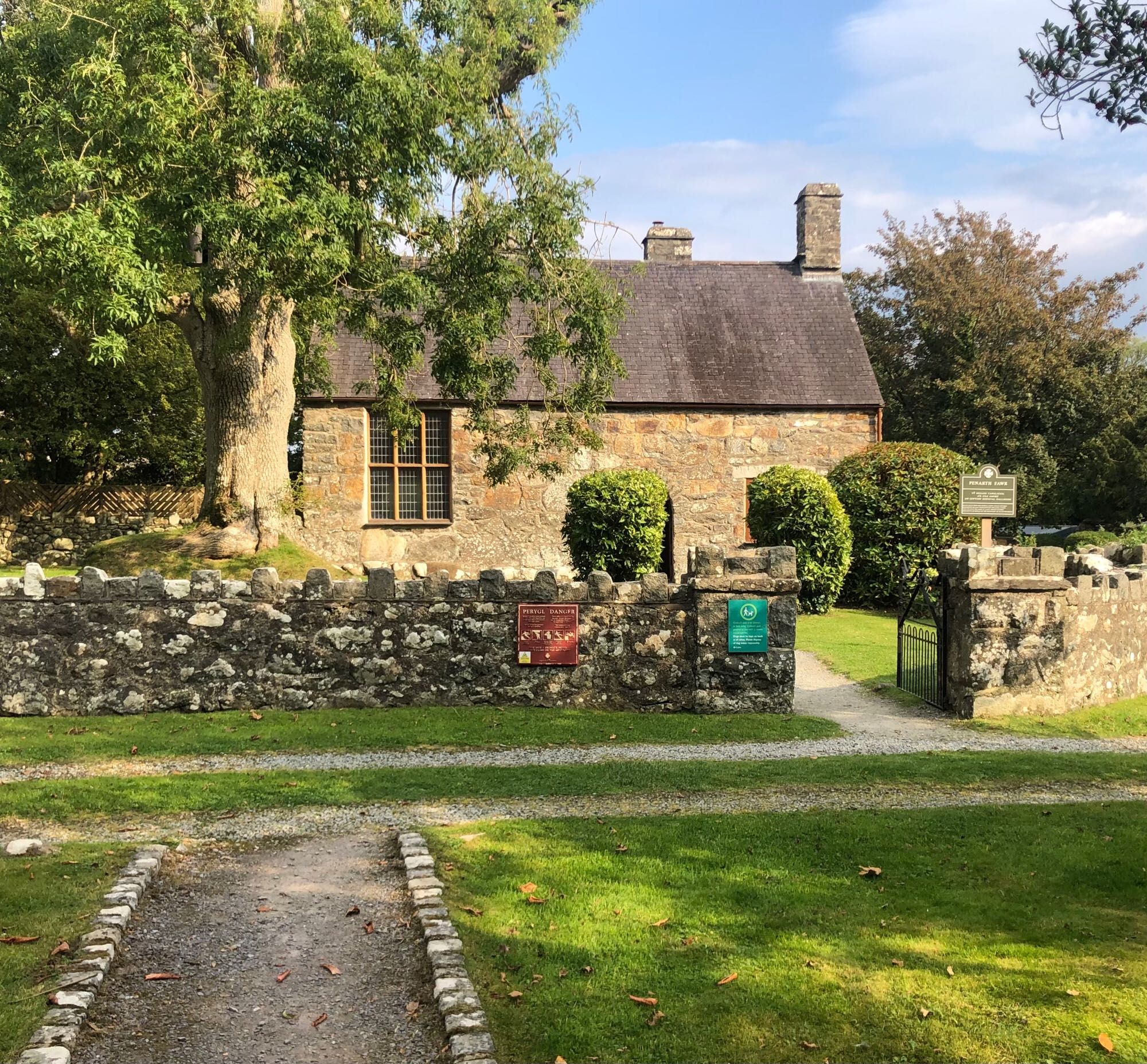 Medieval stone building with large window facing the front, surrounded by trees and shrubs and stone wall around the garden.