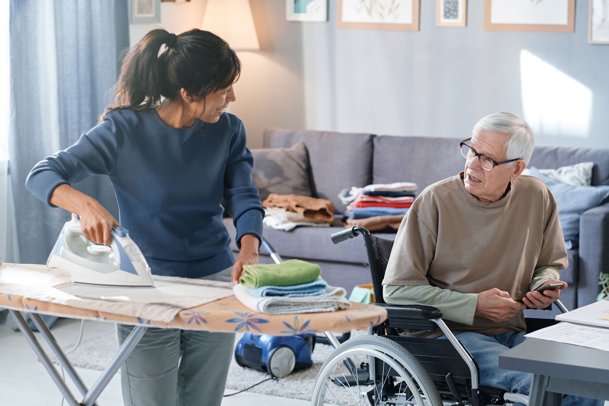 Im Bild: Eine Frau bügelt Kleidung auf dem Bügelbrett in einer Wohnung und spricht mit einem älteren Mann, der im Rollstuhl sitzt. Foto: Anna Stills – Shutterstock.