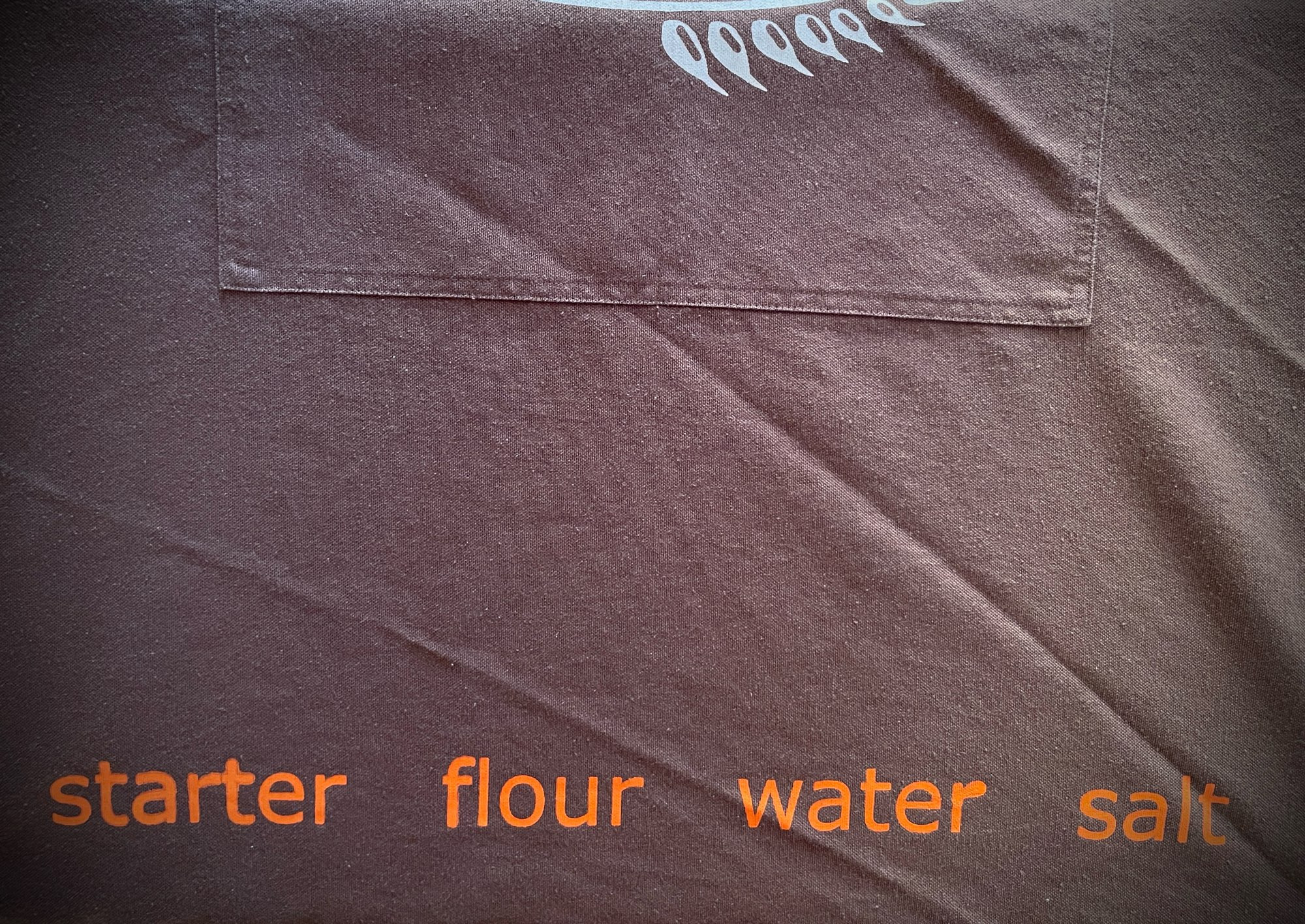 The bottom hem of a brown apron with printing that reads, “starter, flour, water, salt”