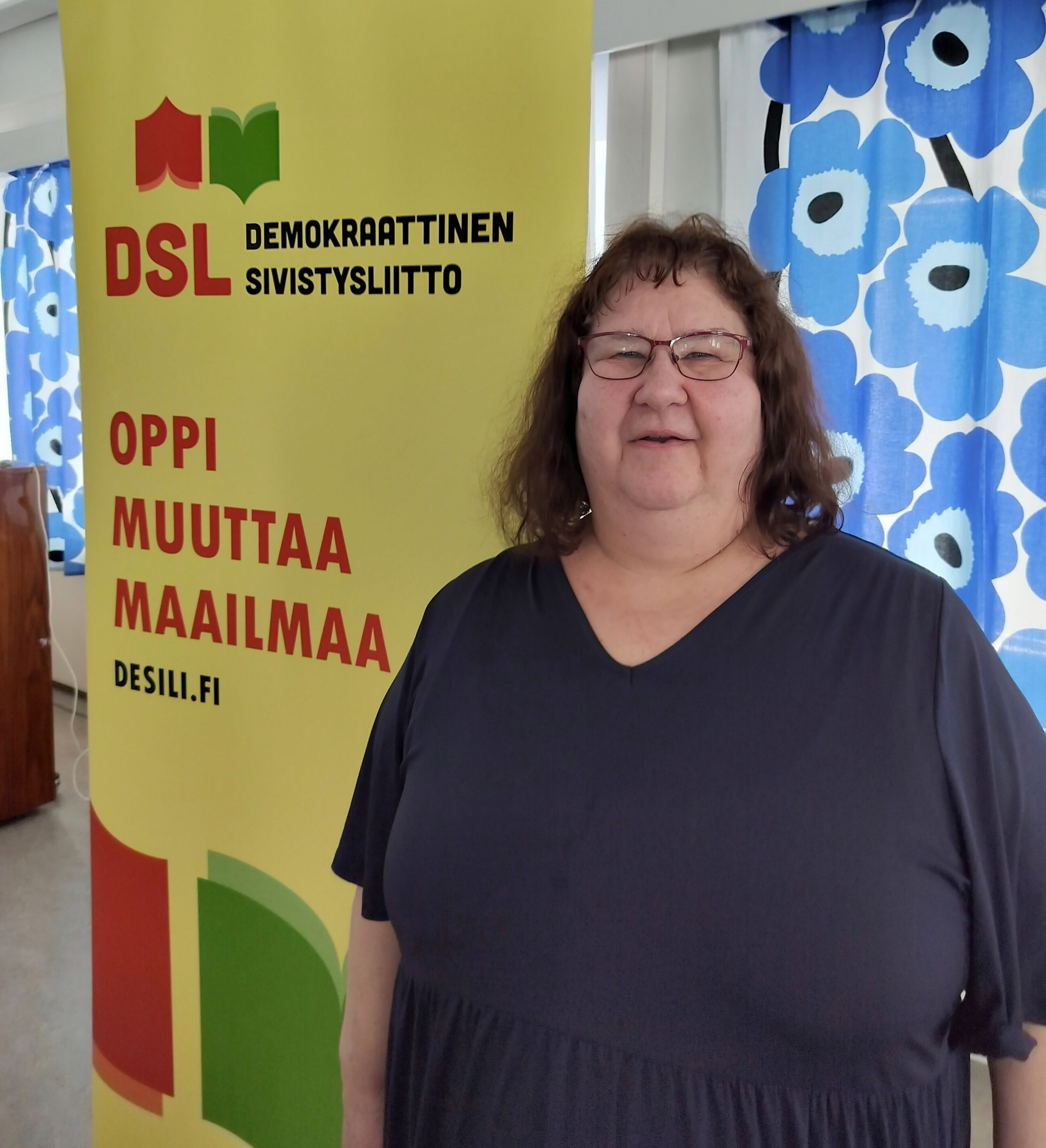 Talvikki Koivu kuvattuna DSL:n uuden roll-upin edessä Sepänkeskuksella, roll-upissa DSL:n logo ja slogan "Oppi muuttaa maailmaa".