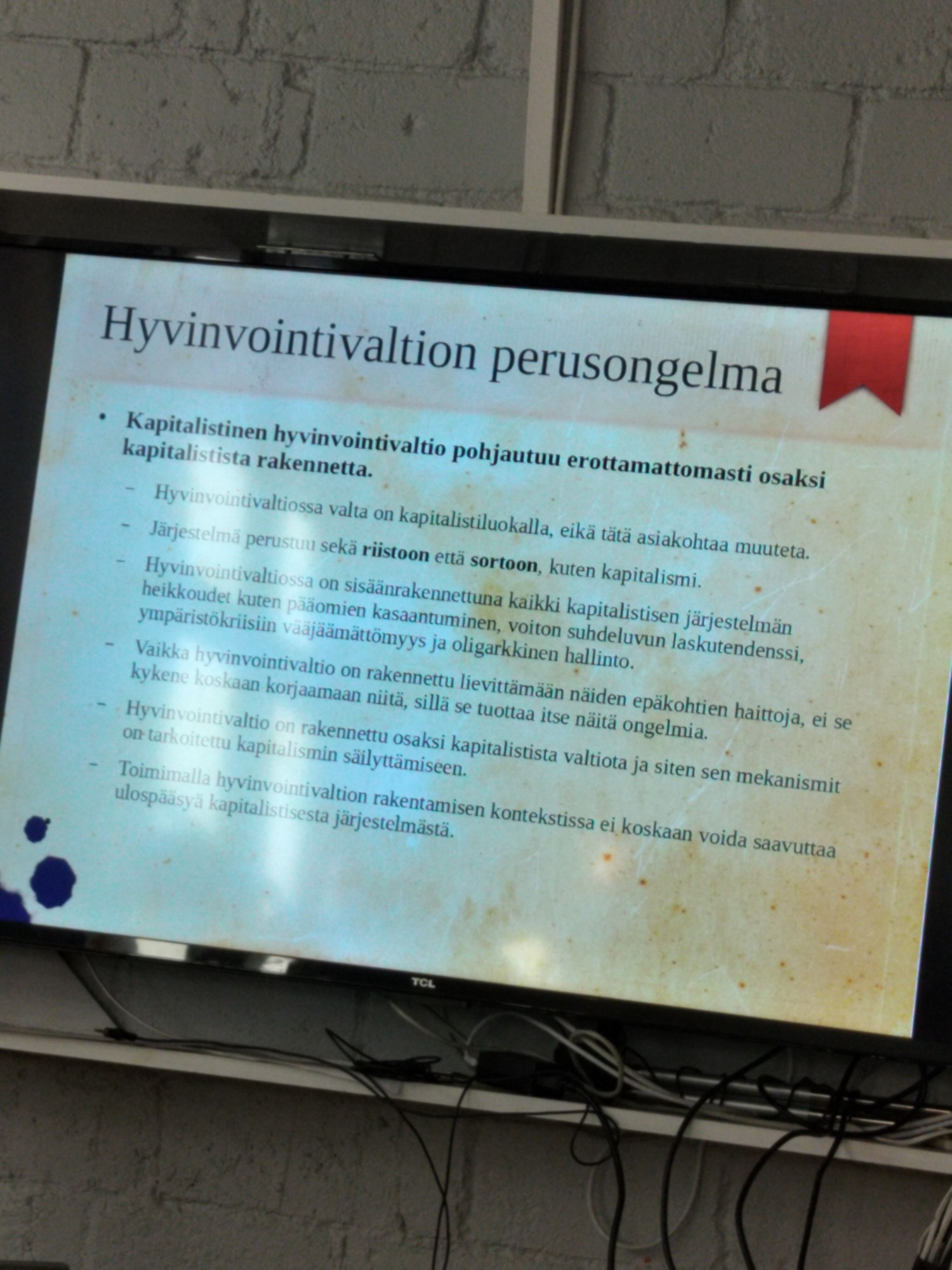 Kuva tv-ruudun näytöltä, jossa alustuksen diaesitys, näkyvillä hyvinvointivaltion perusongelmaa osana kapitalistista järjestelmää.