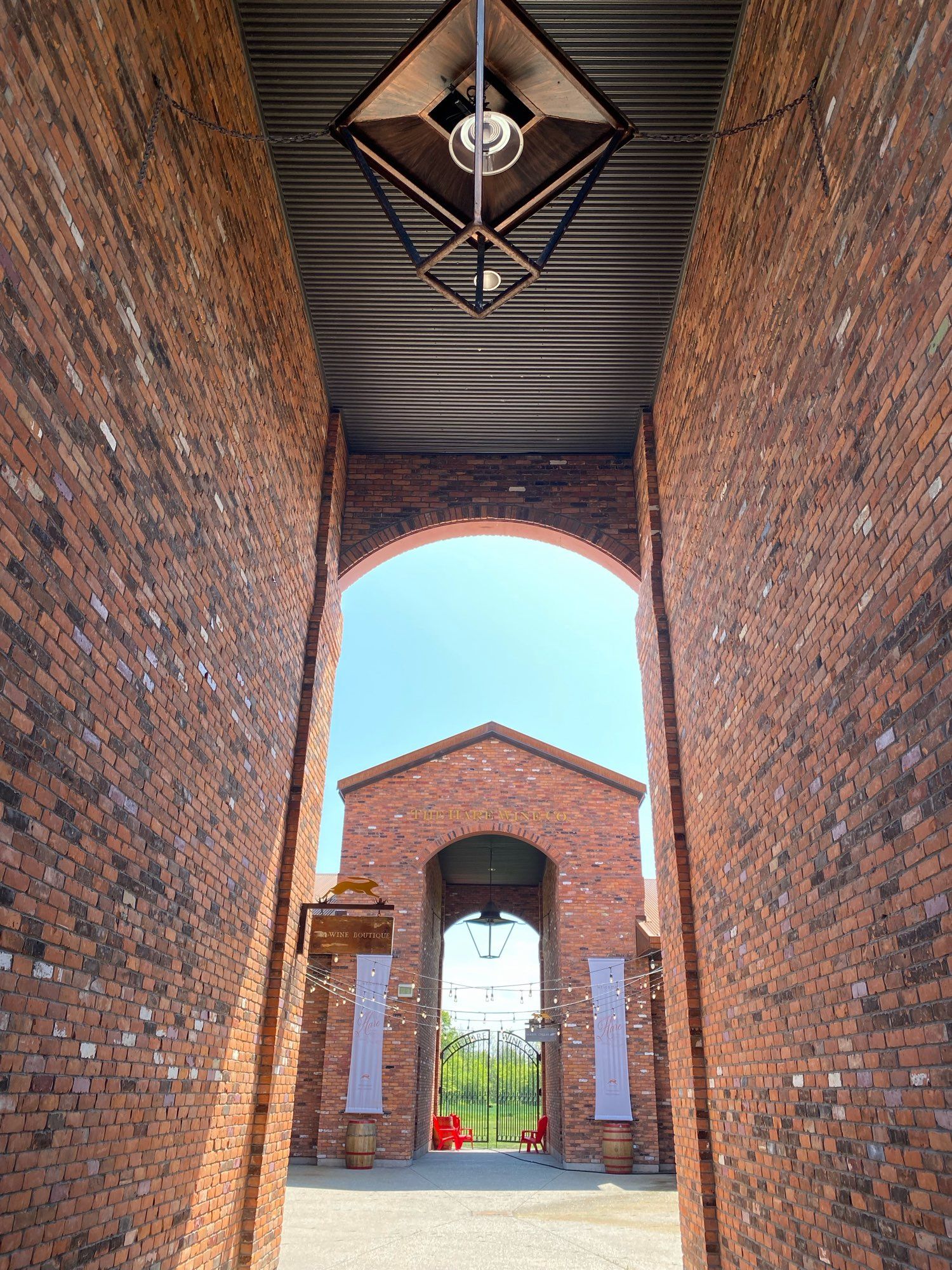 An entry to an exterior courtyard, with two tall brick walls on your left and right. At the far end of the courtyard is a tall archway exit with two more brick walls, echoing the first.