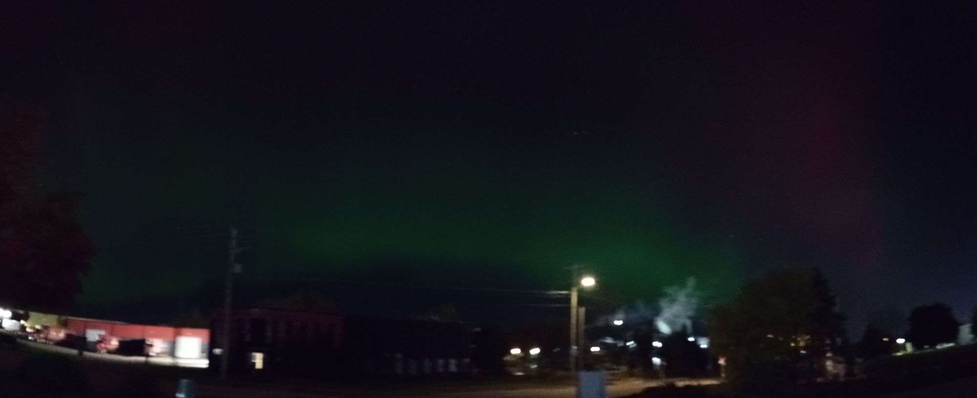 Panoramic photo of aurora borealis flare up, in shades of emerald green. Taken from across the street, in front of a factory.