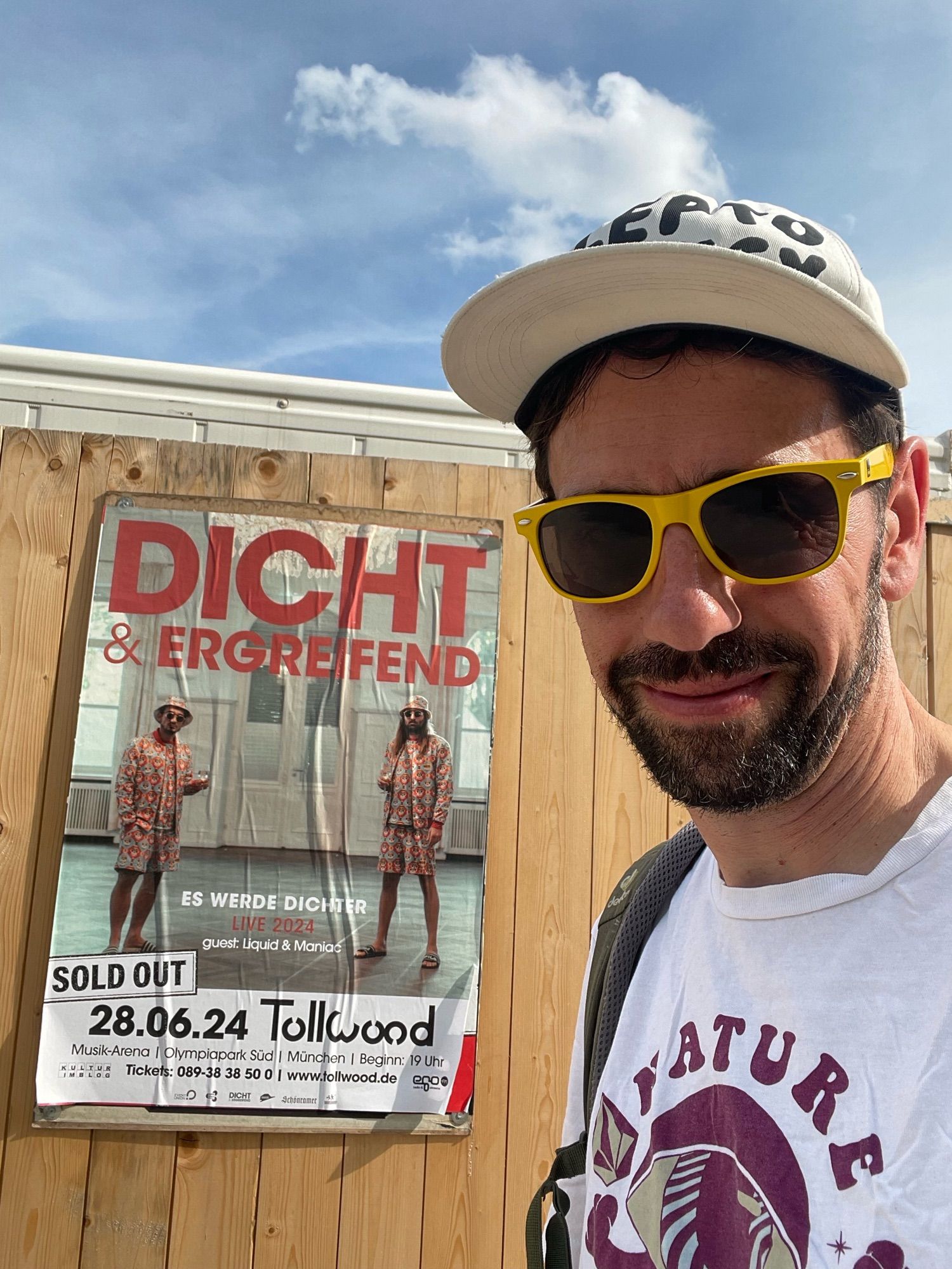 Ein Mann mit Bart und gelber Sonnenbrille steht vor einem Konzertplakat von Dicht & Ergreifend. Sold out 28.06.2024 Tollwood