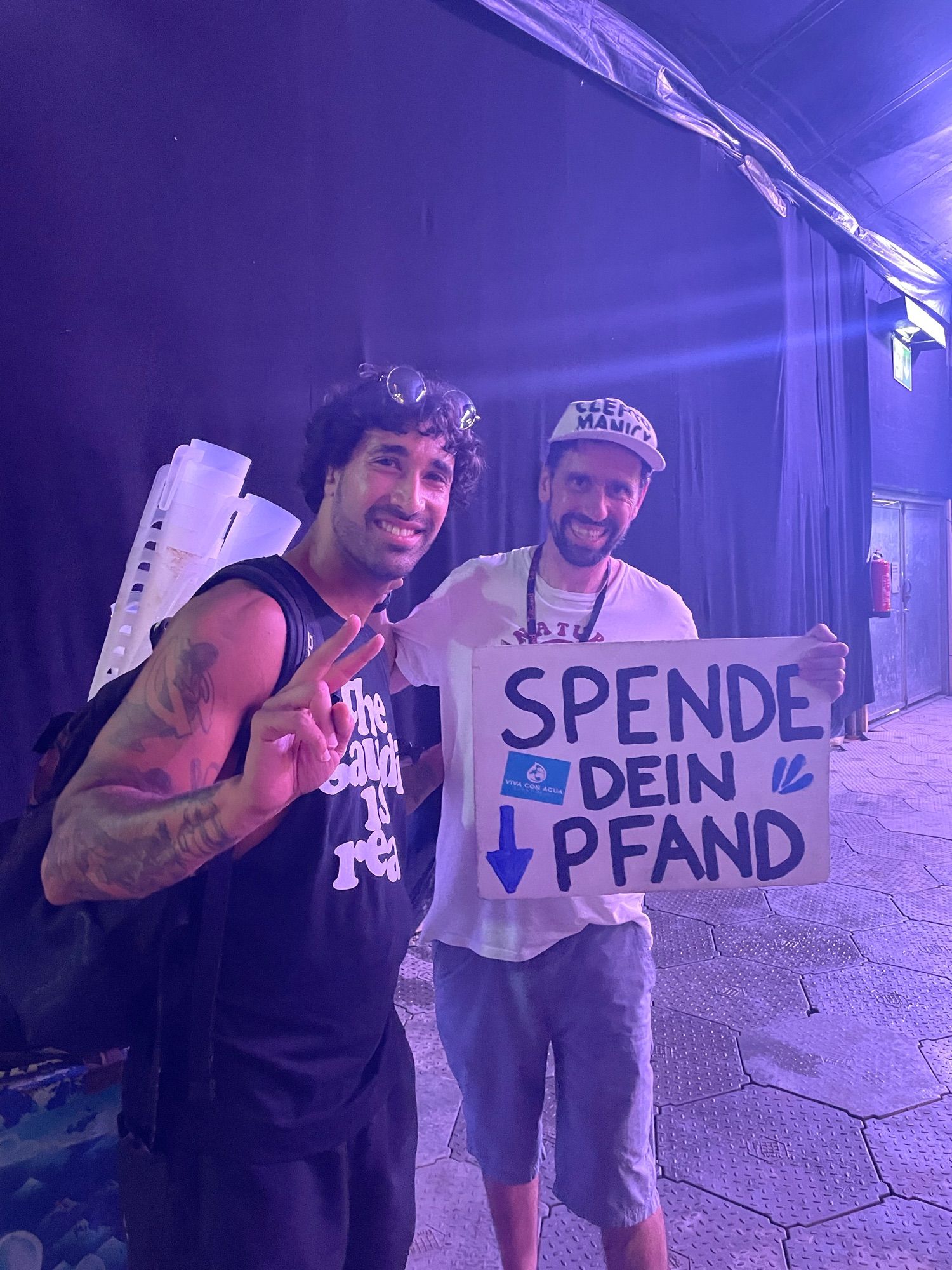 Zwei Personen lächeln in die Kamera. Einer hält ein Schild mit der Aufschrift Spende dein Pfand