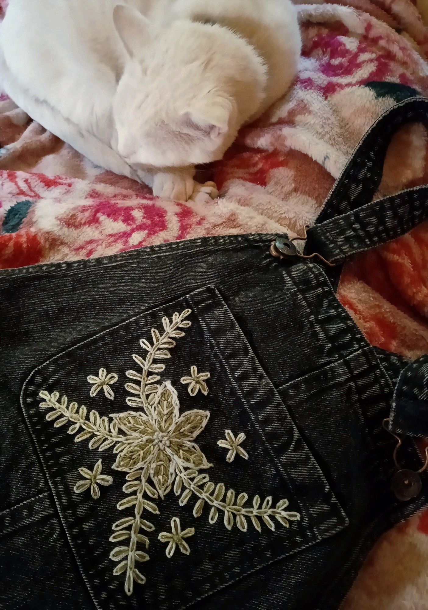 Finished embroidery project: a faded black pair of dungarees lies next to a white sleeping cat oba red floral blanket. The front chest pocket is embroidered with leaves and flowers in white and green thread. The motif is simplistic folk art made with chain stitch.