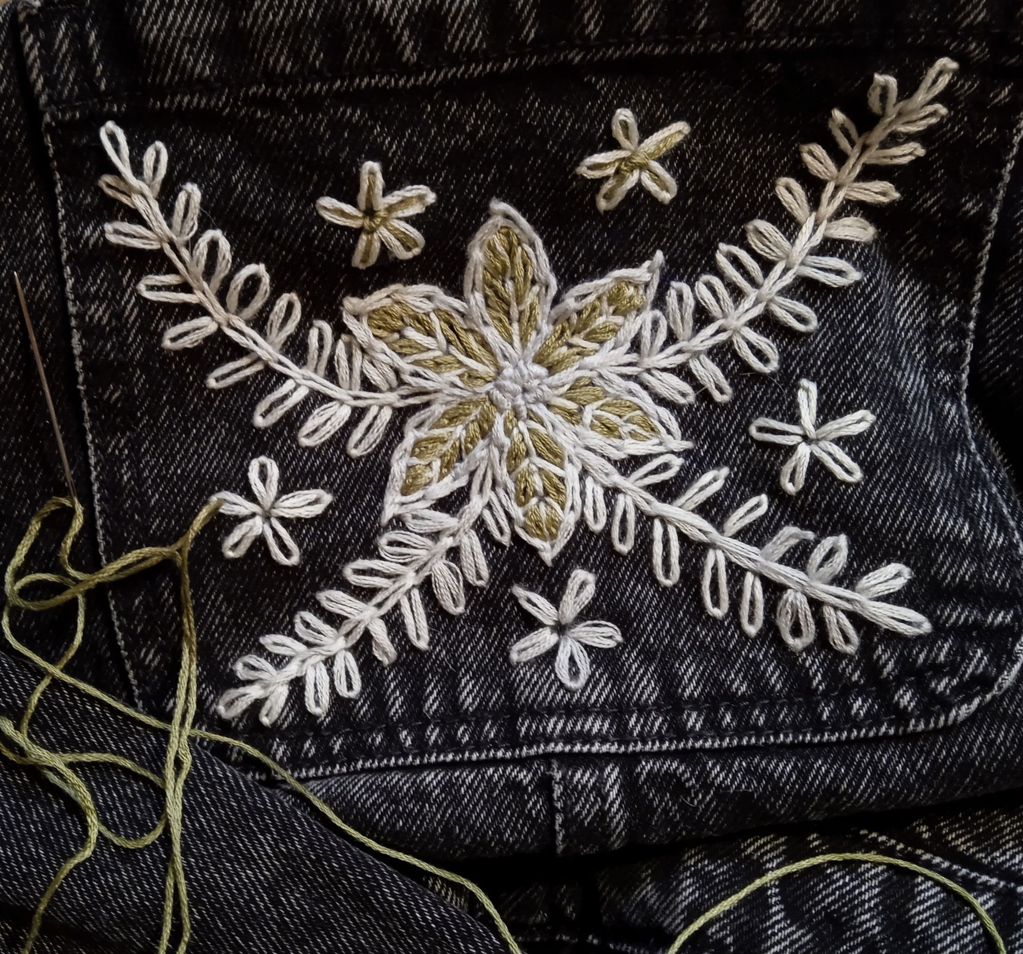 A close up of green and white embroidery on faded black denim. A simplistic flower is stitched in white thread, onto a square pocket. The central flower has 6 petals, and is surrounded by 5 smaller flowers.  4 leafy branches stretch to the corners. Loose green thread and a needle lie below the pocket on the folds of denim.