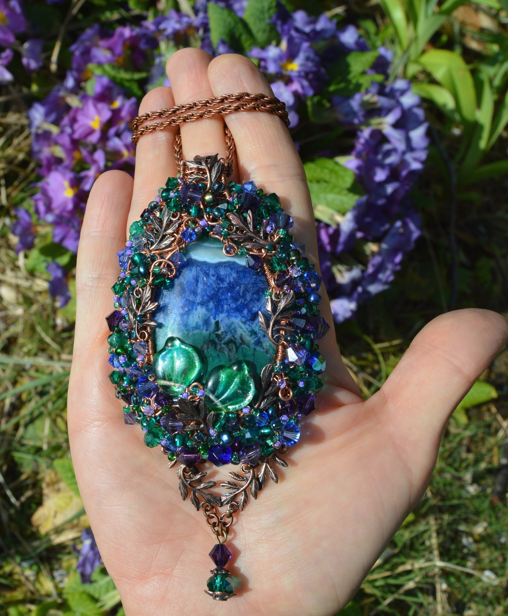 A vivid blue purple and green pendant necklace worked with beads and tiny copper fern leaves. Displayed on an open hand, with blooming purple primroses in the background. The pendant is teardrop shaped, and fills the palm - about 3 inches tall. The central stone is marbled dark blue and green, and surrounded by a frame of woven copper wire into which tiny crystals and beads are worked. below hand 2 tiny leaves and a purple and green beaded pendant.