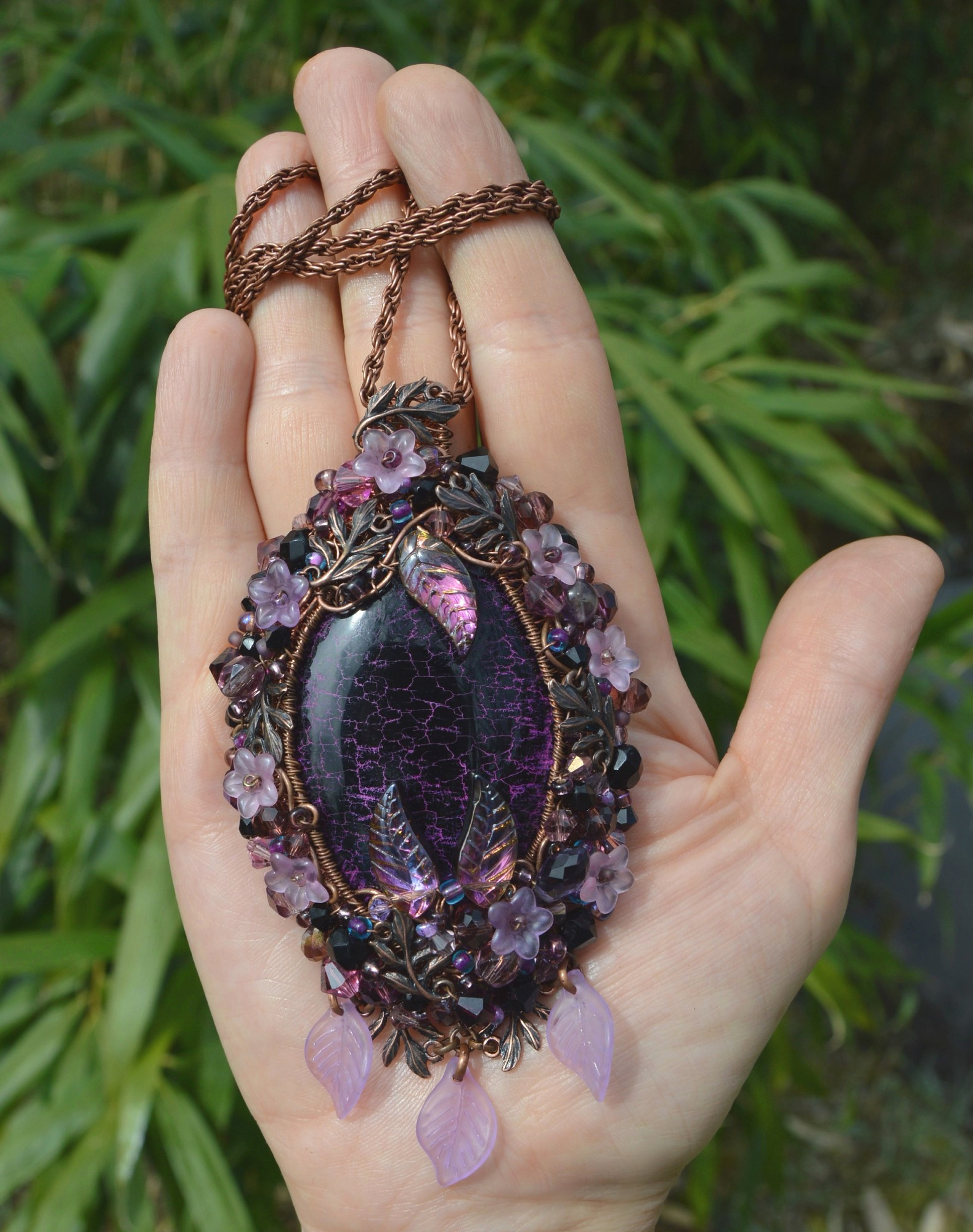 A black and purple beaded pendant, lying in the palm of a hand against a background of lush green leaves. Oval shaped and 3 inches long, the central stone is black with pinkish cracks. A frame of copper wirewrapping is sewn with flower beads, pink and purple crystals and beads, and tiny fern leaf charms, 3 pink peaves hang underneath.