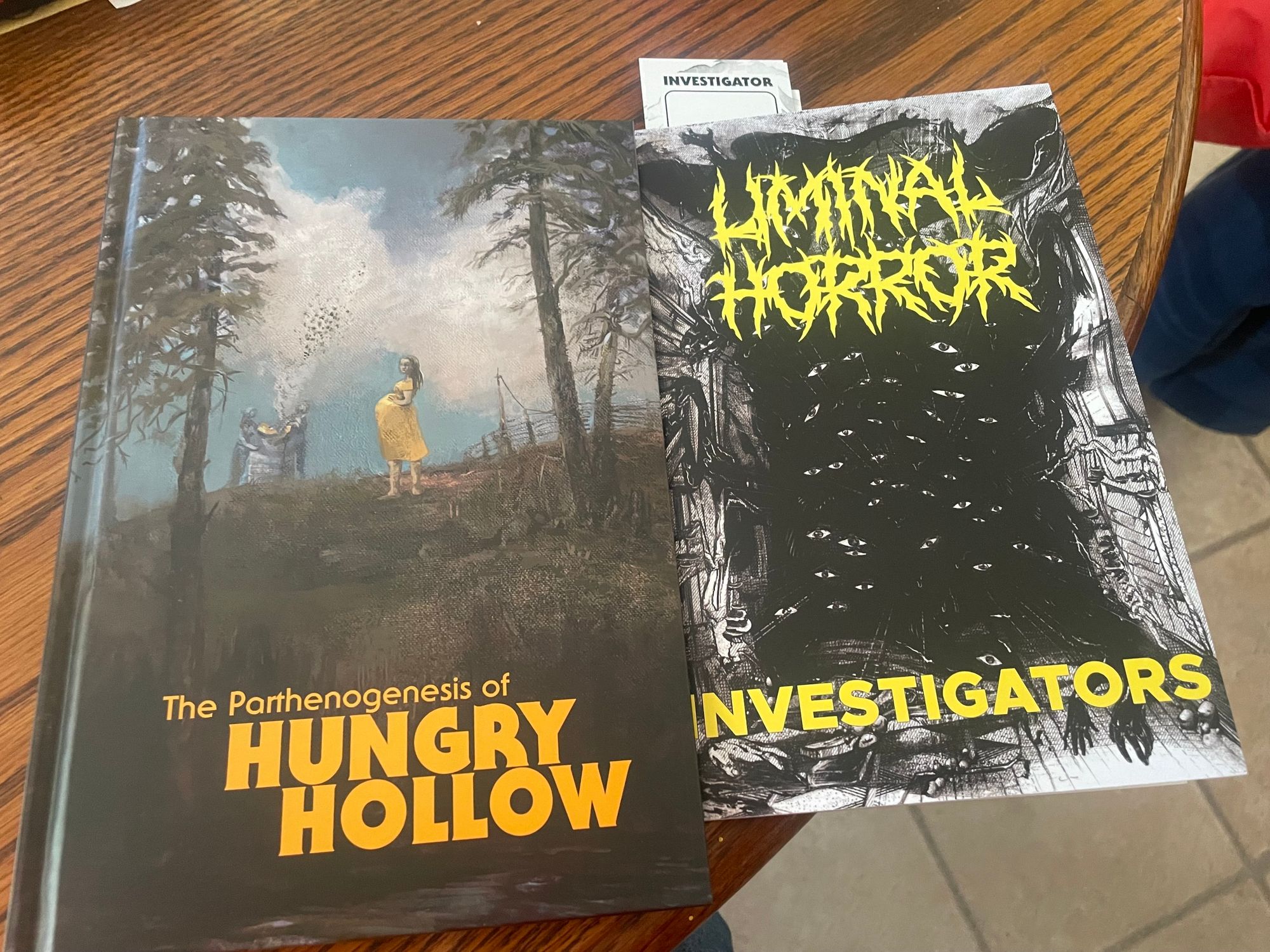 The image depicts two books on a wooden table. On the left is "The Parthenogenesis of Hungry Hollow," featuring a colorful cover illustration of a pregnant woman in a yellow dress near two beekeepers. On the right is "Liminal Horror: Investigators