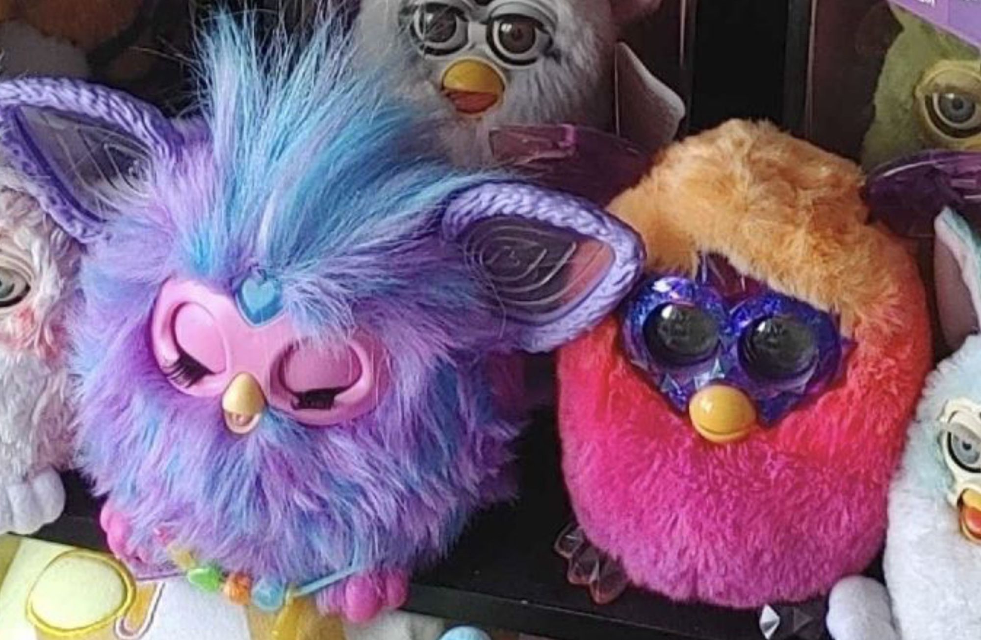 A very furry Furby with pastel fur in blue and mauve with a pink mask and pink feet, eyes closed in apparent bliss, next to a short-furred Furby with a purple-sparkle mask and big dark eyes, fur is the colours of the Lesbian Pride flag