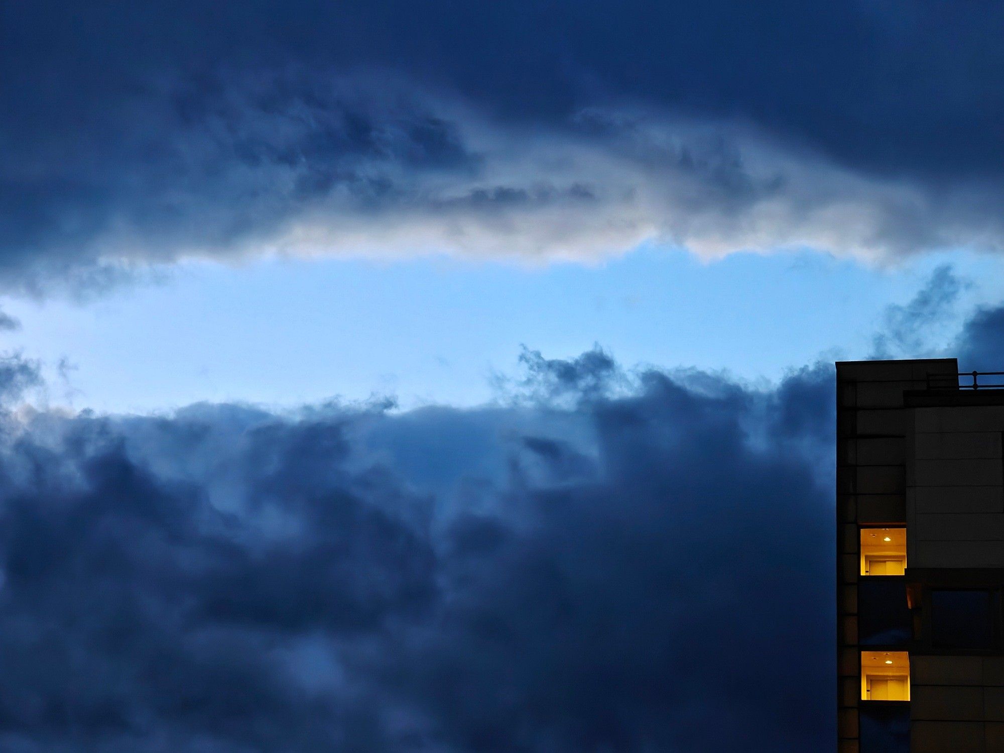 Evening view of a corner of a modern building with two lit windows, in front of dark clouds, with a wide hole through which the blue sky and some sun lit cloud edge are visible.