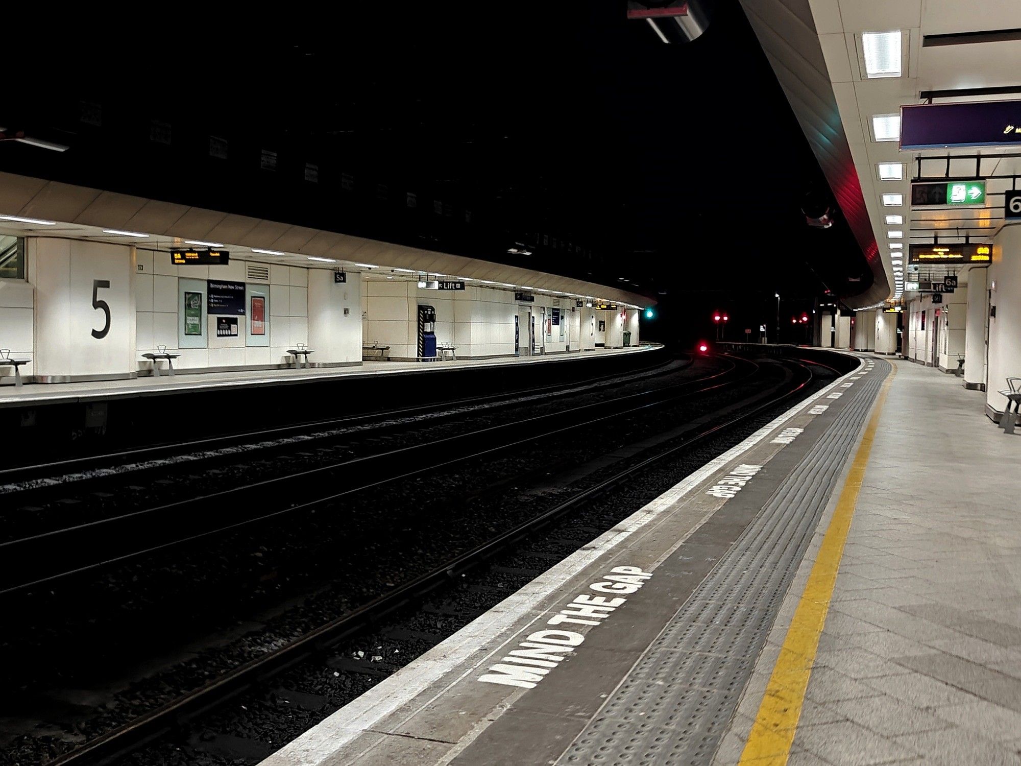 A deserted train station somewhere in the UK. The tracks are dark, practically black, whereas the platforms are brightly lit and mostly white. A yellow line runs the length of the platform. 'Mind the gap' is repeated in white letters along the platform edge.