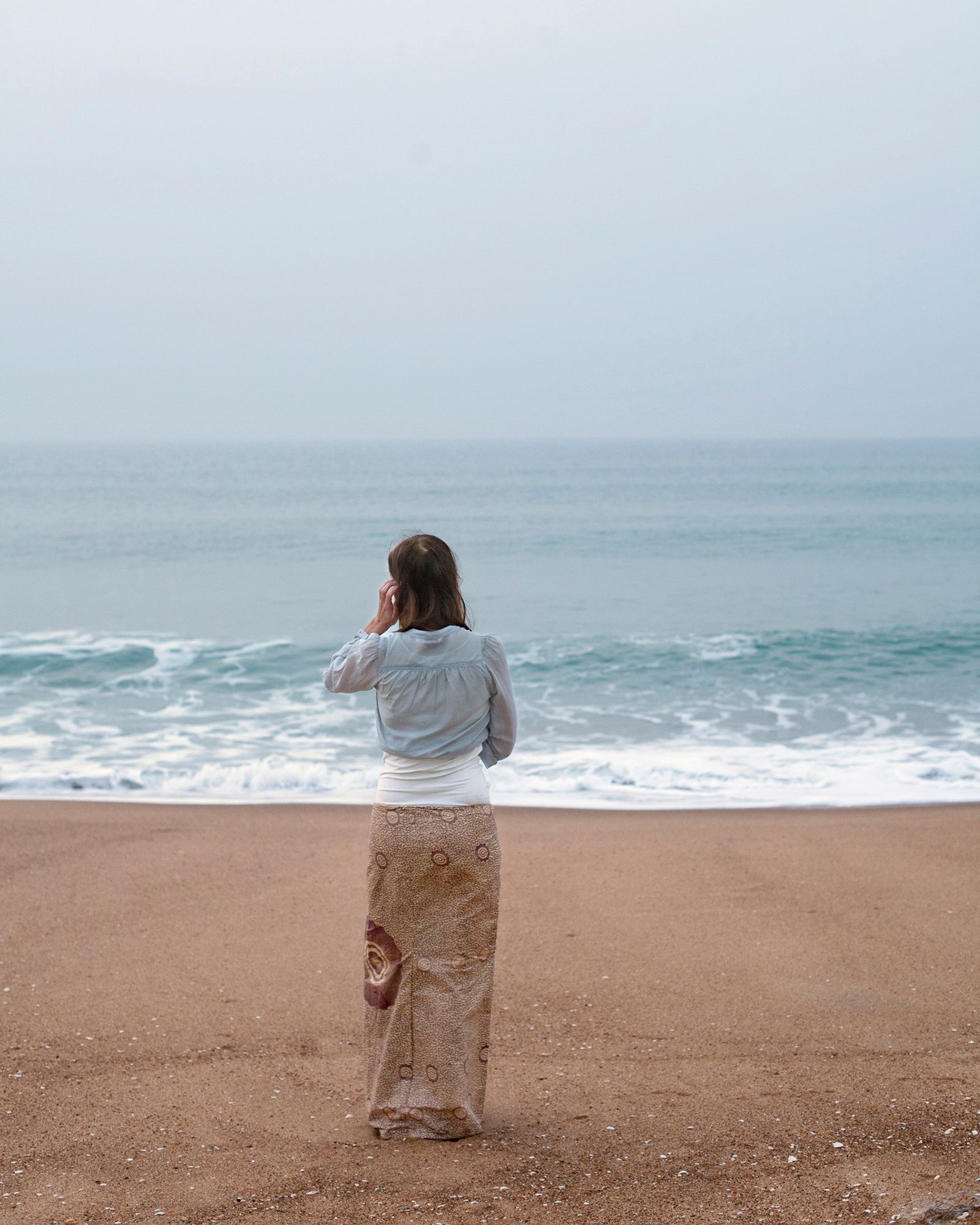 Photo by Wilma Hurskainen. A person standing on a sandy beach looking out over the sea. The person's skirt and top match and are aligned with the colors of the beige beach, the white surf and the blue of the sea.