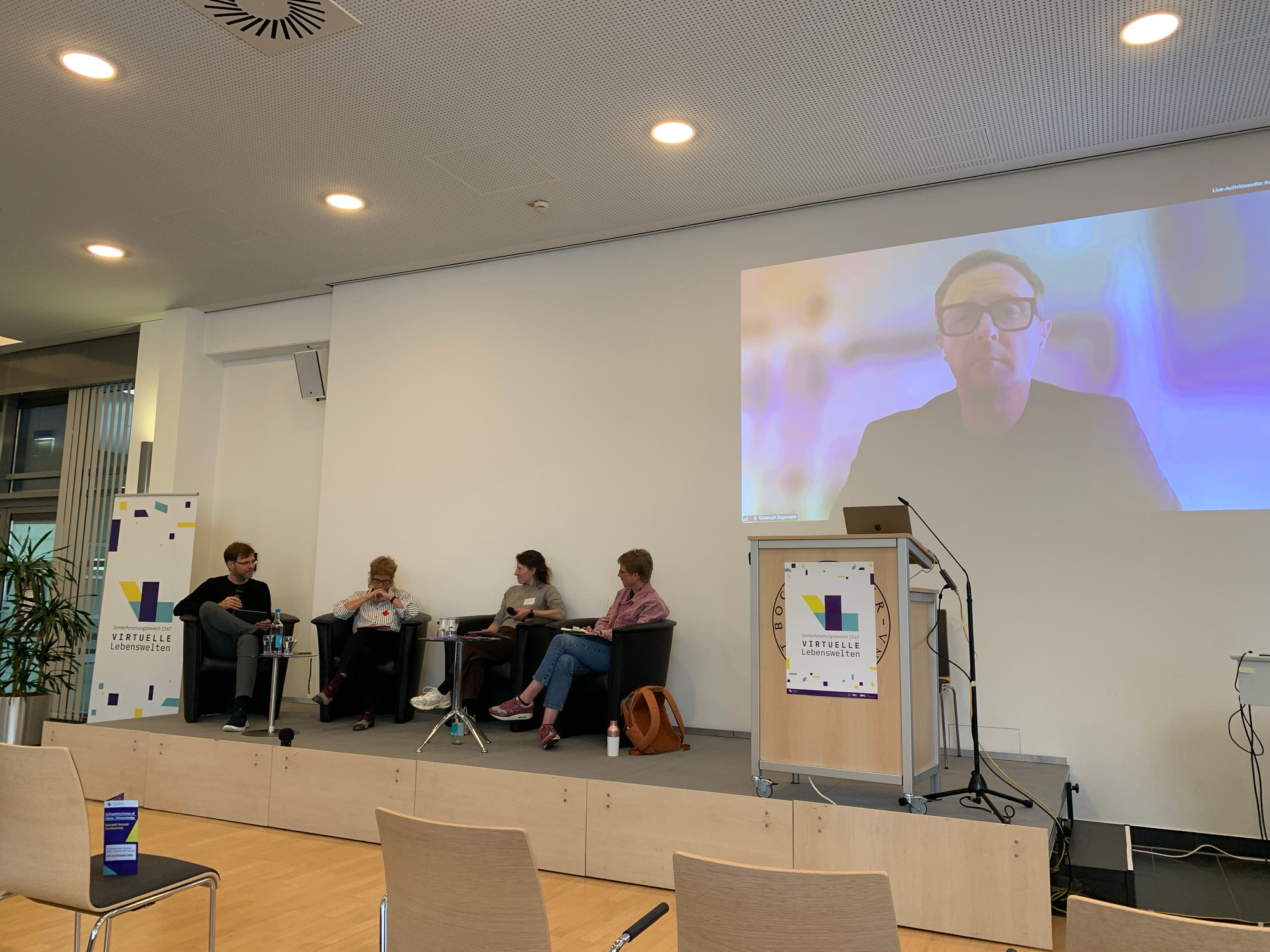 Panel of 4 people in chairs on a stage, plus one on a big Zoom screen: Florian Sprenger (moderator), Anne Dippel, Patrizia Breil, Iris van der Tuin, plus Christoph Engemann (on Zoom)