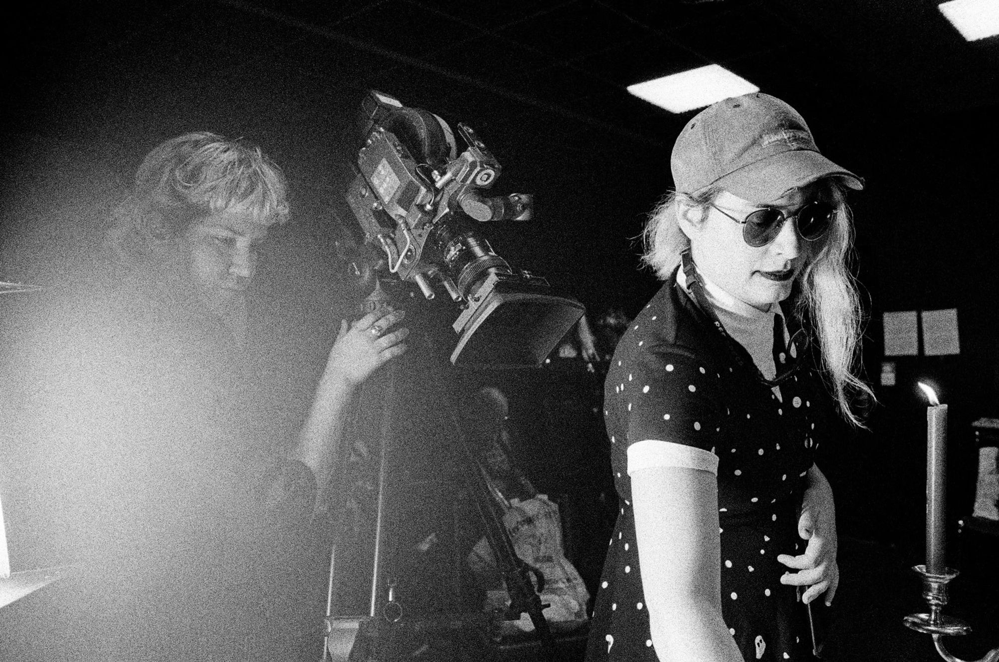 On a set, Morgann is fixing something on a table, behind her her trusted first AC, he is holding Molly an Arri SR3, a big 16mm camera. Behind him an actor is getting ready in the shadows.