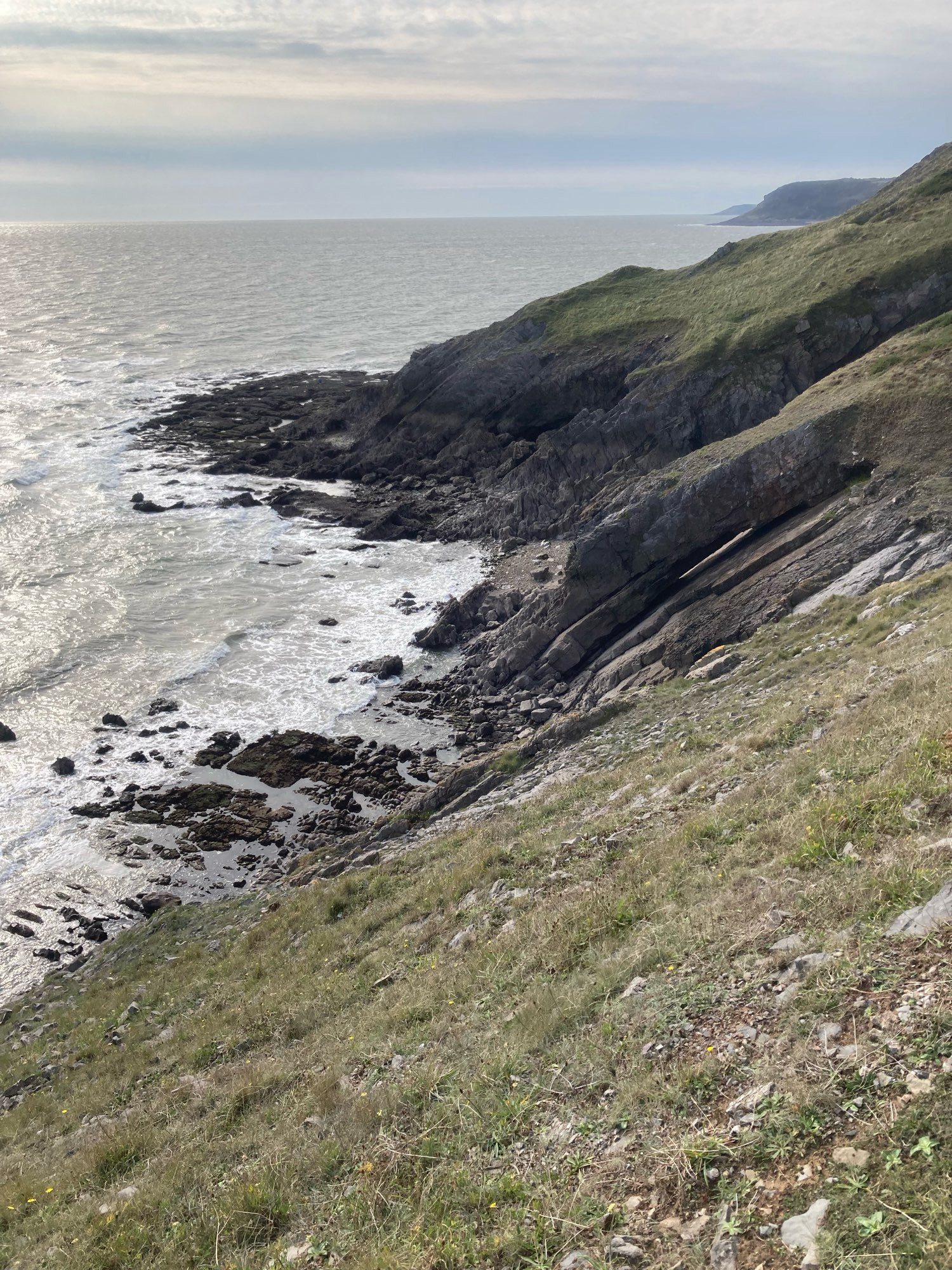 View from a cliff top path down to a rocky coast below. The sea is relatively calm and the sky streaked with clouds.