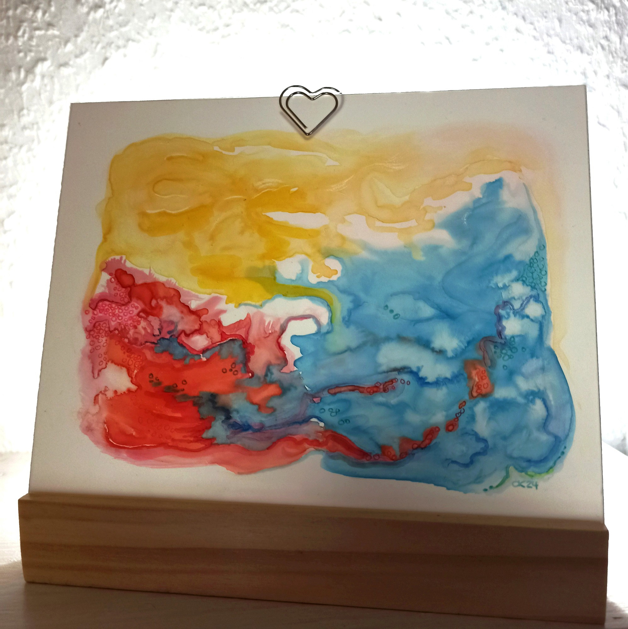 Von hinten beleuchteter Bilderrahmen aus zwei Plexiglasscheiben, die in einem Vierkantholz stecken.
Darin Aquarell auf Synthetikpapier in Gelb, Rot und Blau. Die wolkigen Flächen verschwimmen ineinander.
Die Plexischeiben werden von einer kleinen Büroklammer in Herzform zusammengehalten.