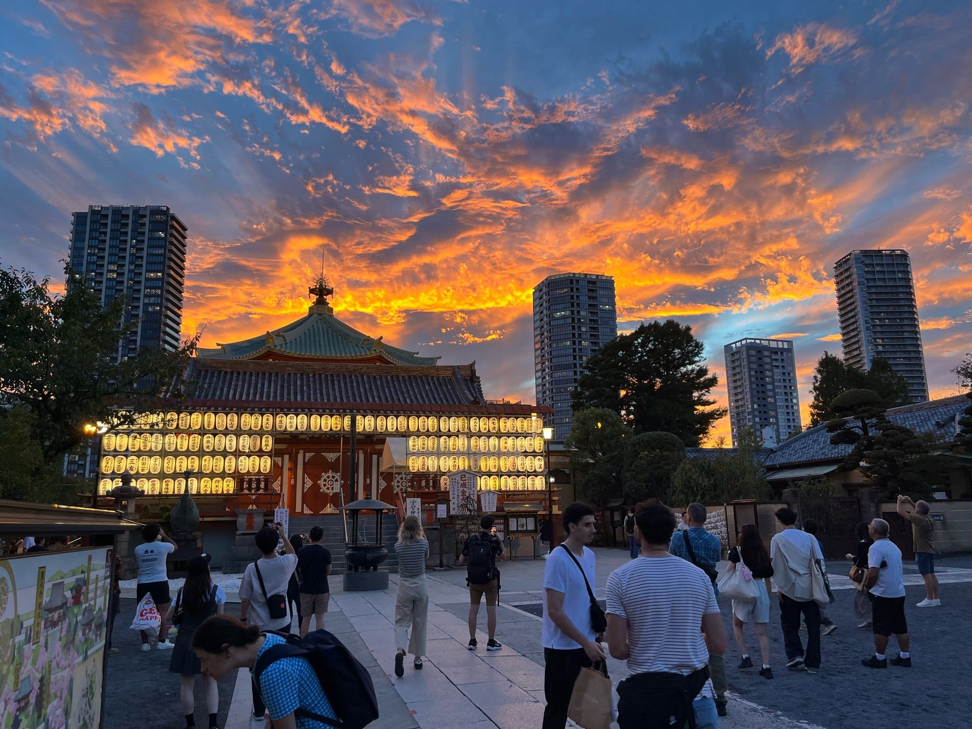 A flamboyant sky with torn clouds over one of the shrines and multiple skyscrapers near the Ueno parc pond. Lots of people going around or taking pictures of the beautiful scenery.