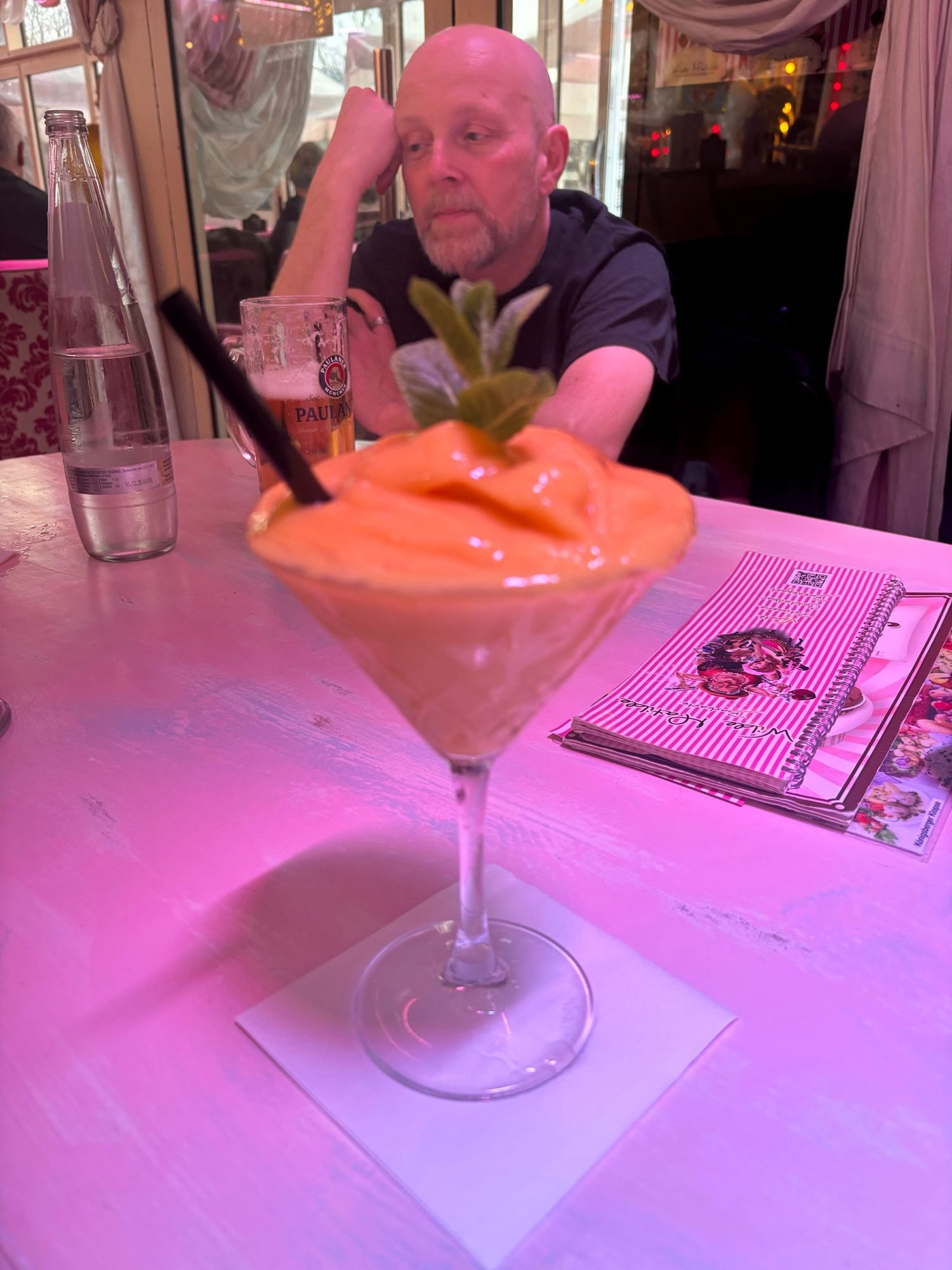 A peach colour led cocktail on the table of the Circu Bar in Berlin; a bald man with a bear considers a glass of beer in front of him at the far side of the table