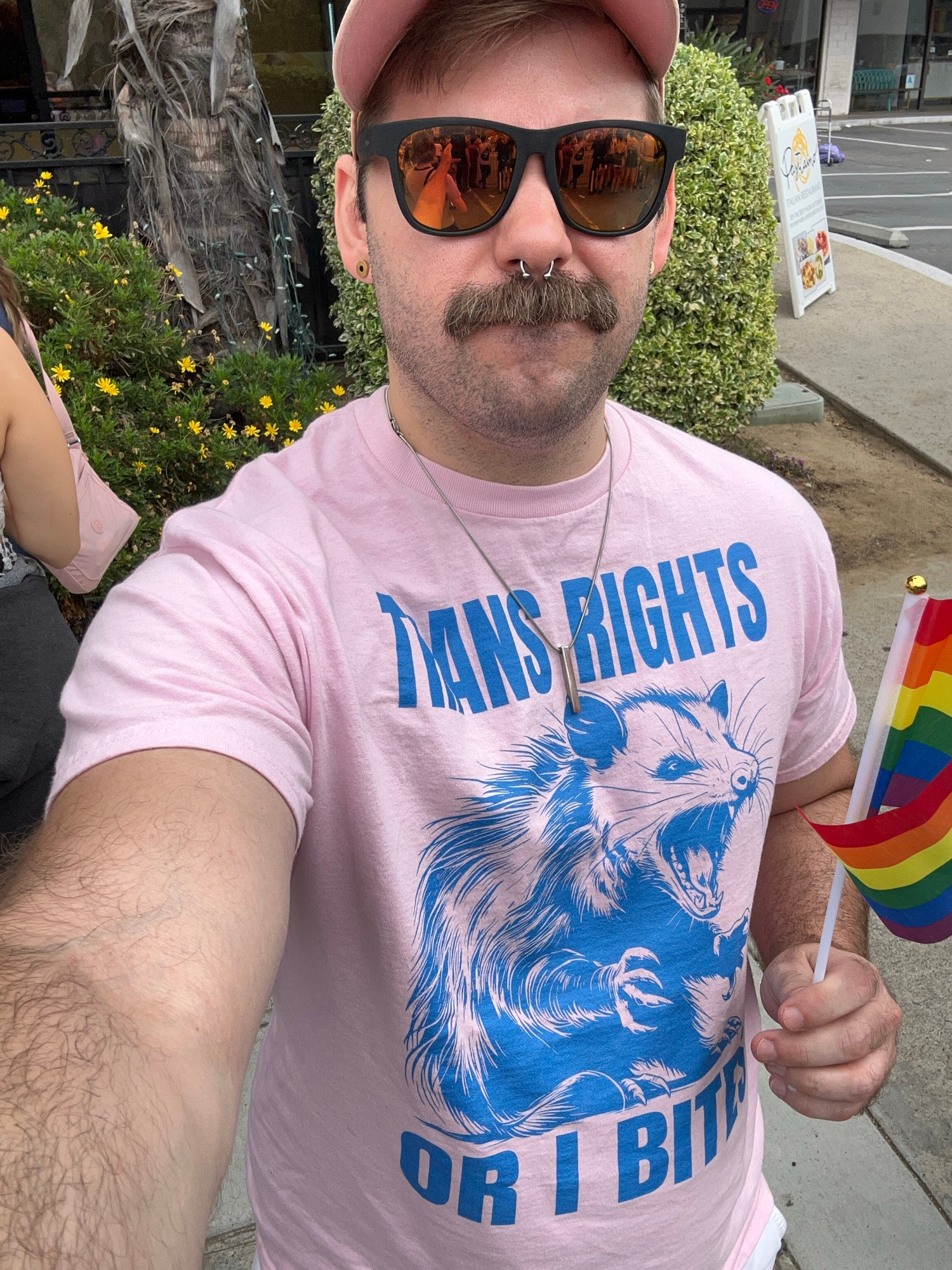 Me wearing a pink shirt with a possum on it. It reads Trans Rights or I Bites. I’m holding a rainbow flag. 