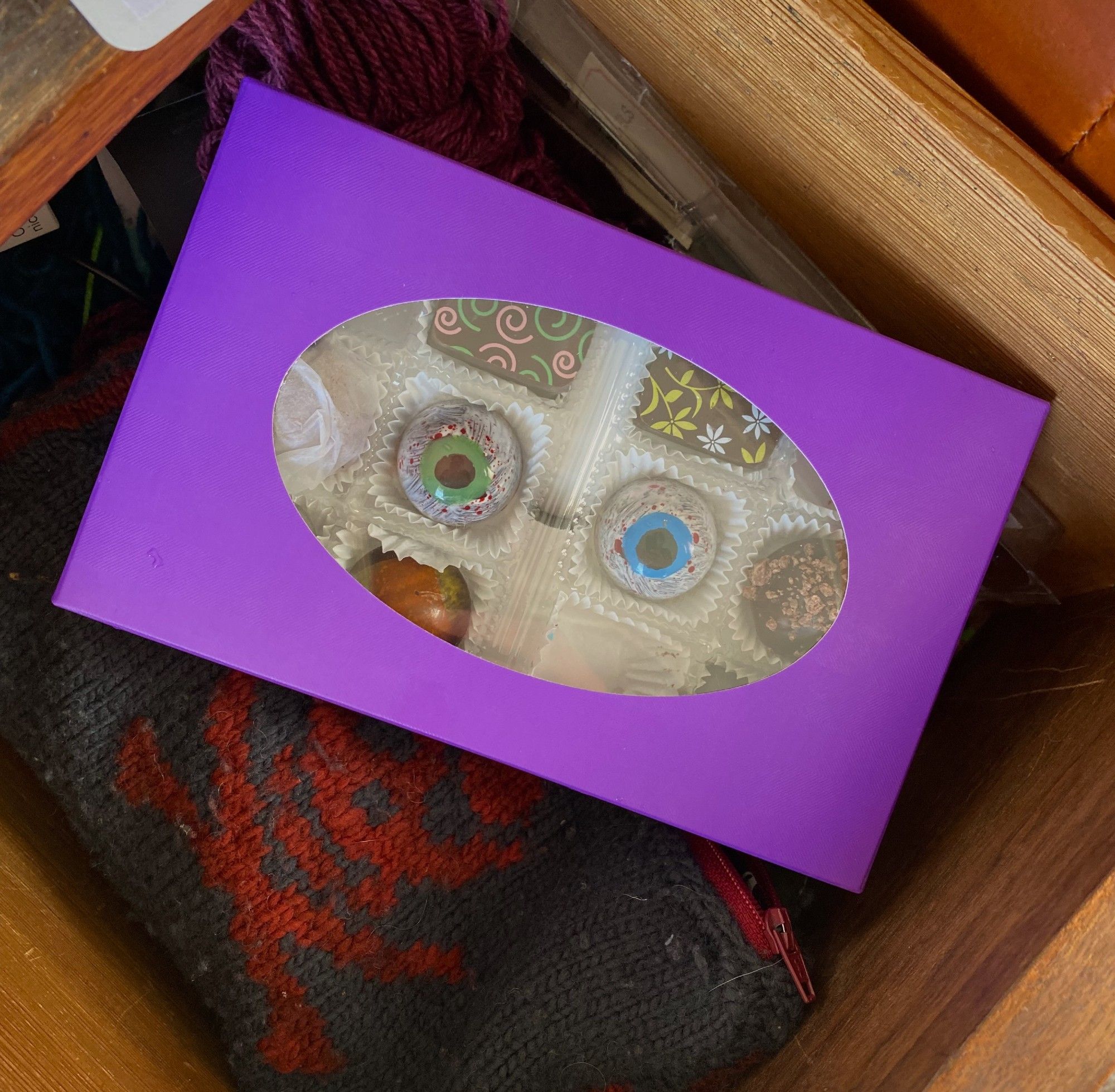 A purple chocolate box with an oval window in the top, sitting in a drawer. A pair of chocolates decorated to look like eyeballs--one blue, one green--stare out from the window of the box.