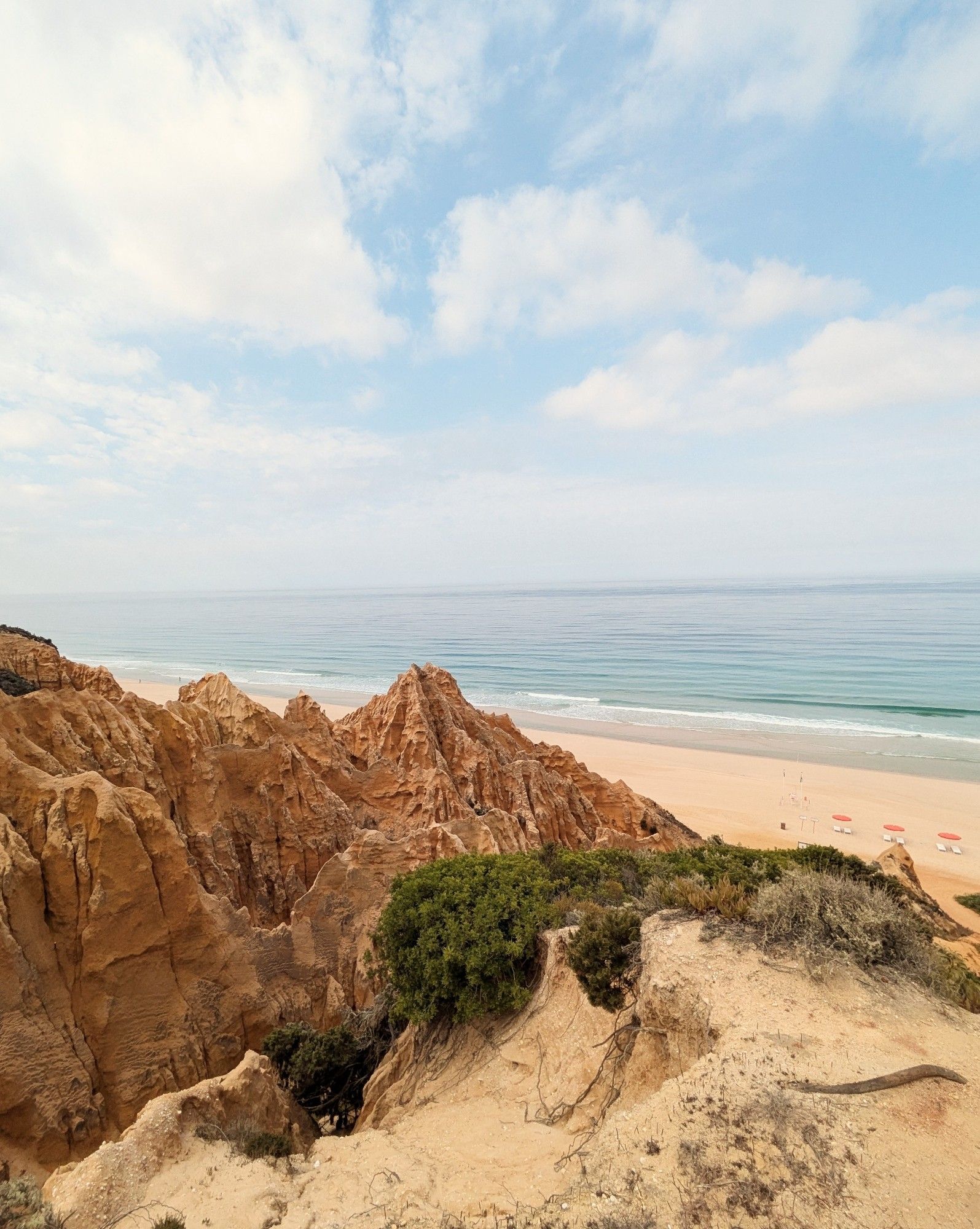 A rocky red cliff juts into a sandy beach. The sky is light blue with white clouds. Melides, Portugal.