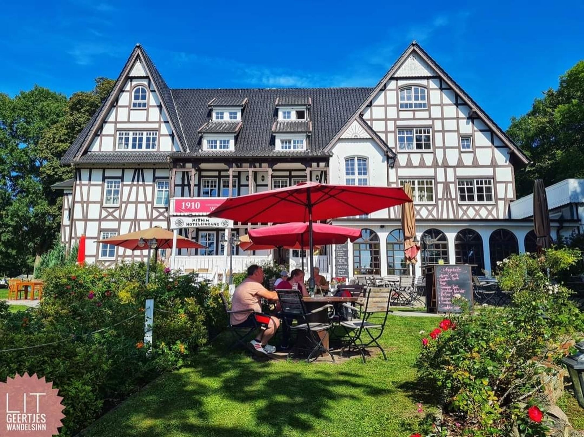 Ein Fachwerkhaus vor blauem Himmel mit roten Sonnenschirmen davor. Leute sitzen darunter. Der Rasen grün, rote Rosen im Vordergrund.